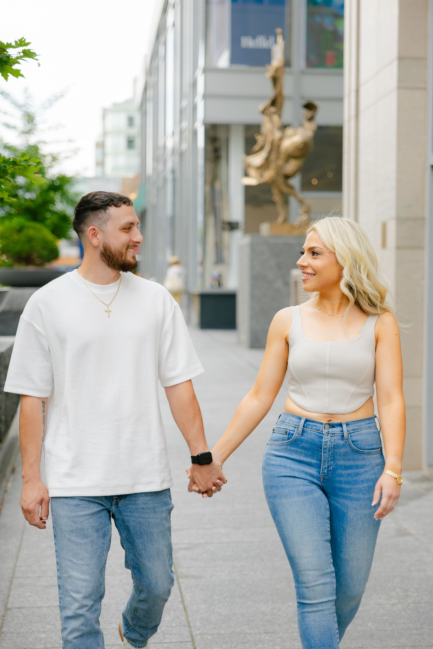 A couple in matching casual outfits walking hand in hand on a city sidewalk near modern buildings.