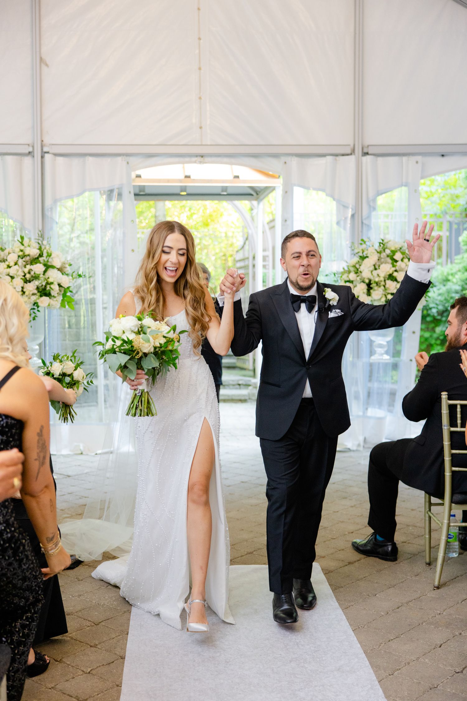 A newlywed couple walks down the aisle in celebration as the bride holds a bouquet and both have their arms raised.