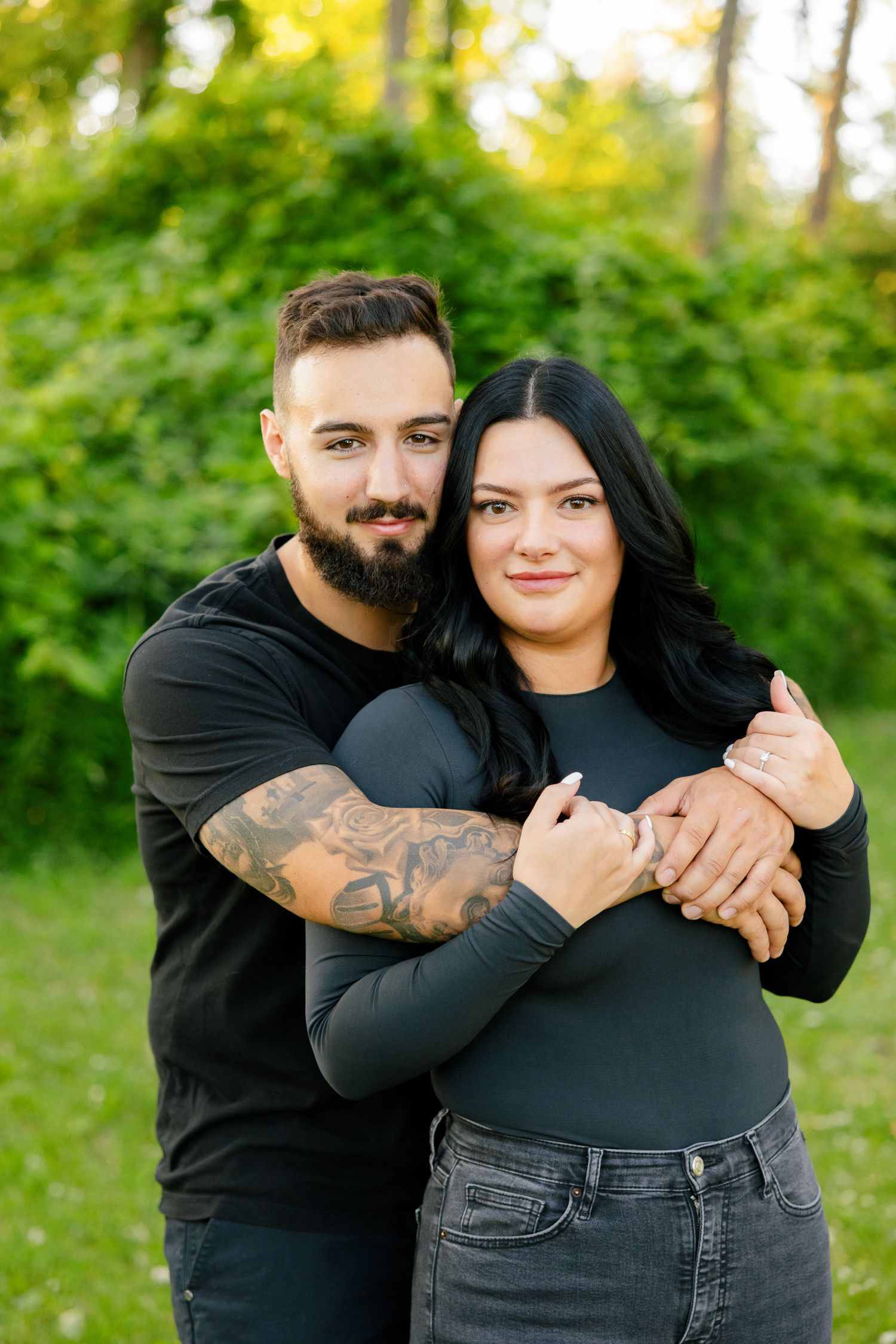 Couple embracing in coordinated black outfits against lush green forest background.