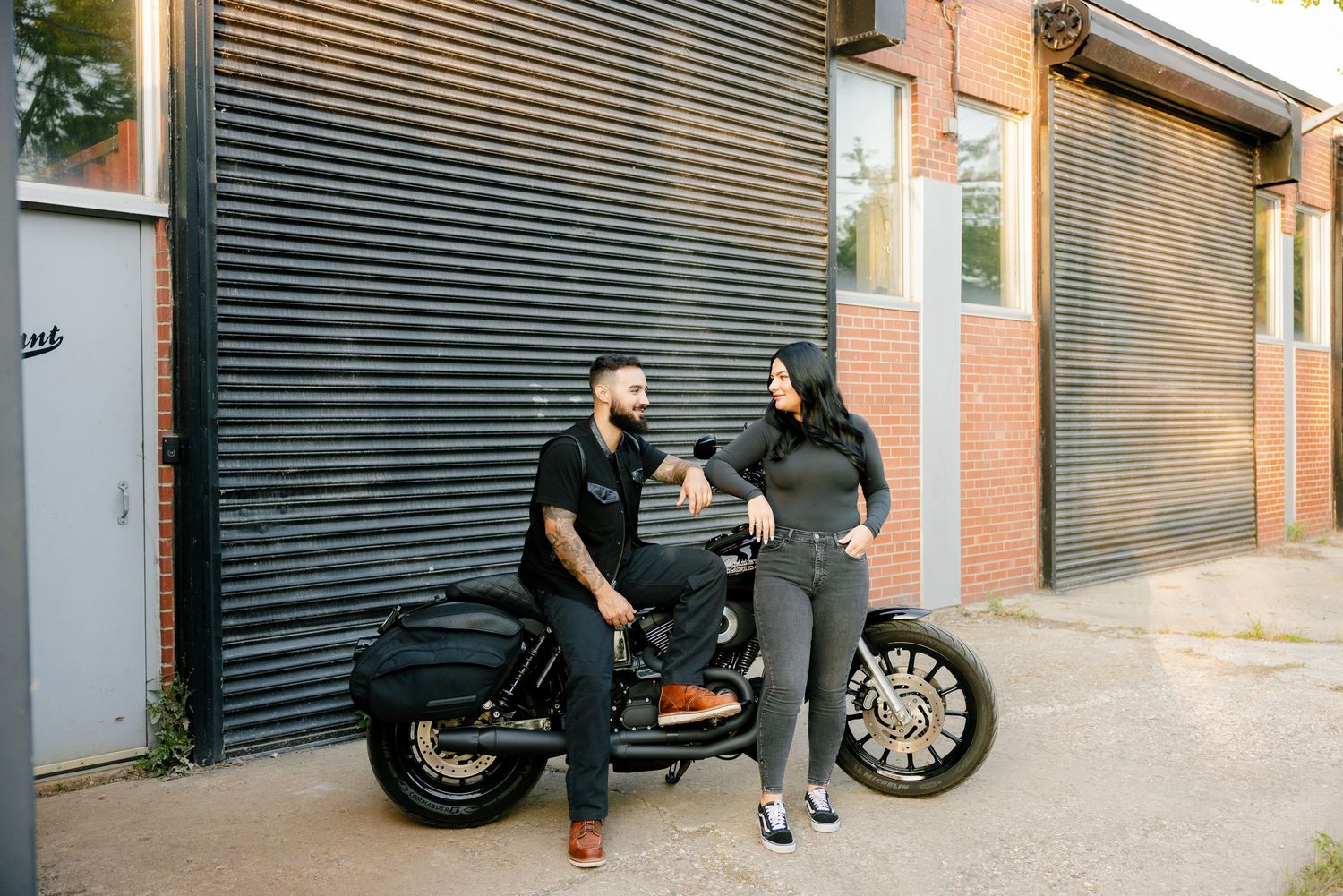 Two people chatting while standing next to a parked motorcycle by a corrugated metal wall.