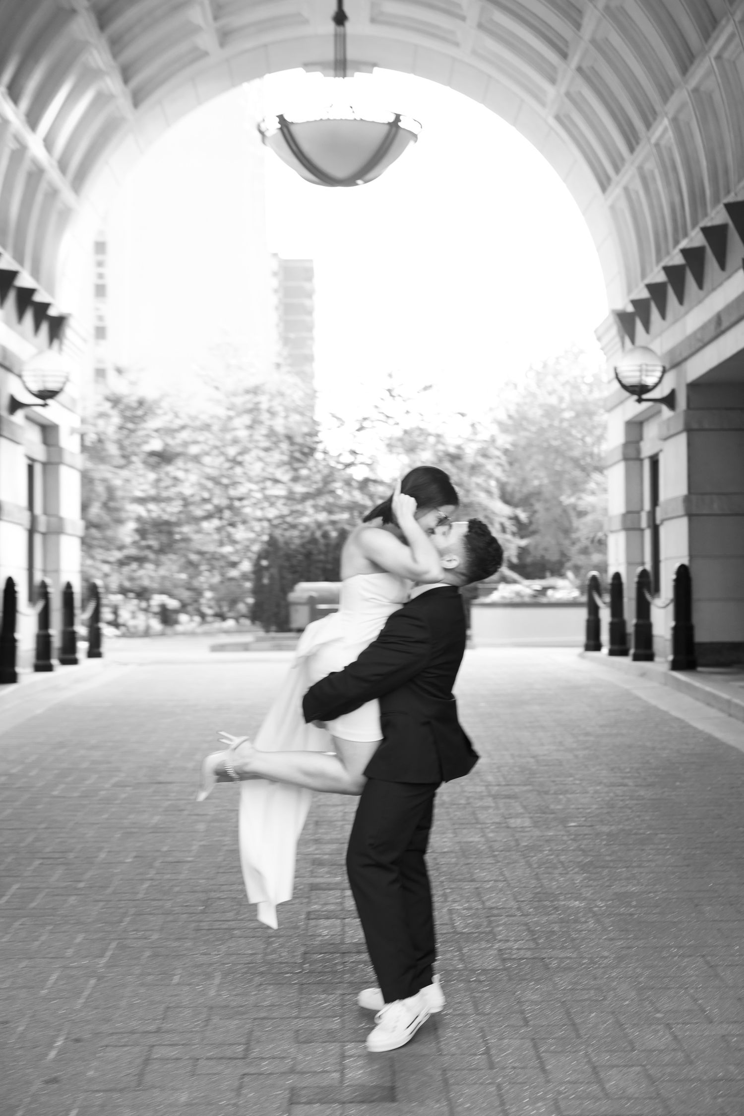 Romantic black and white photo of a couple in formal wear sharing a playful embrace under an archway.