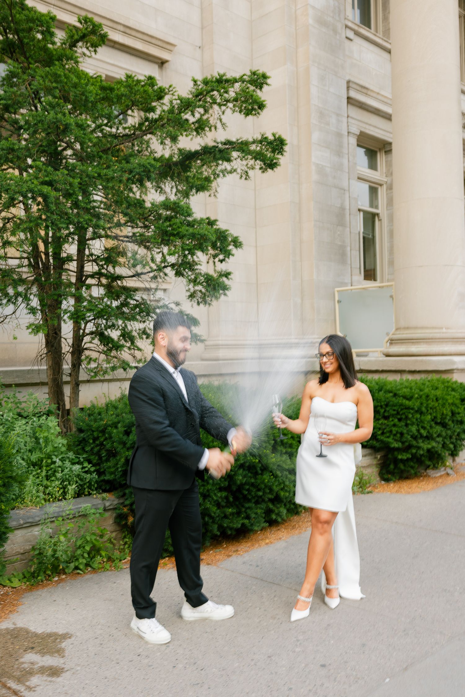 A couple celebrates with champagne spray outside a stately building with green foliage.