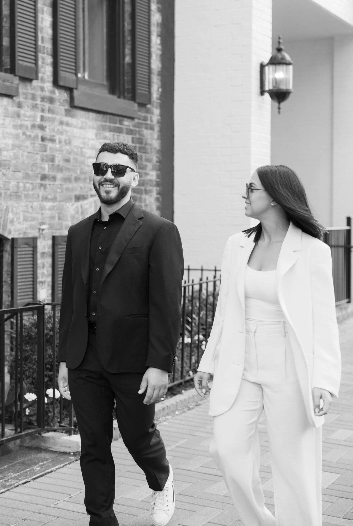 A black and white photo of a couple in contrasting outfits walking along a brick building.