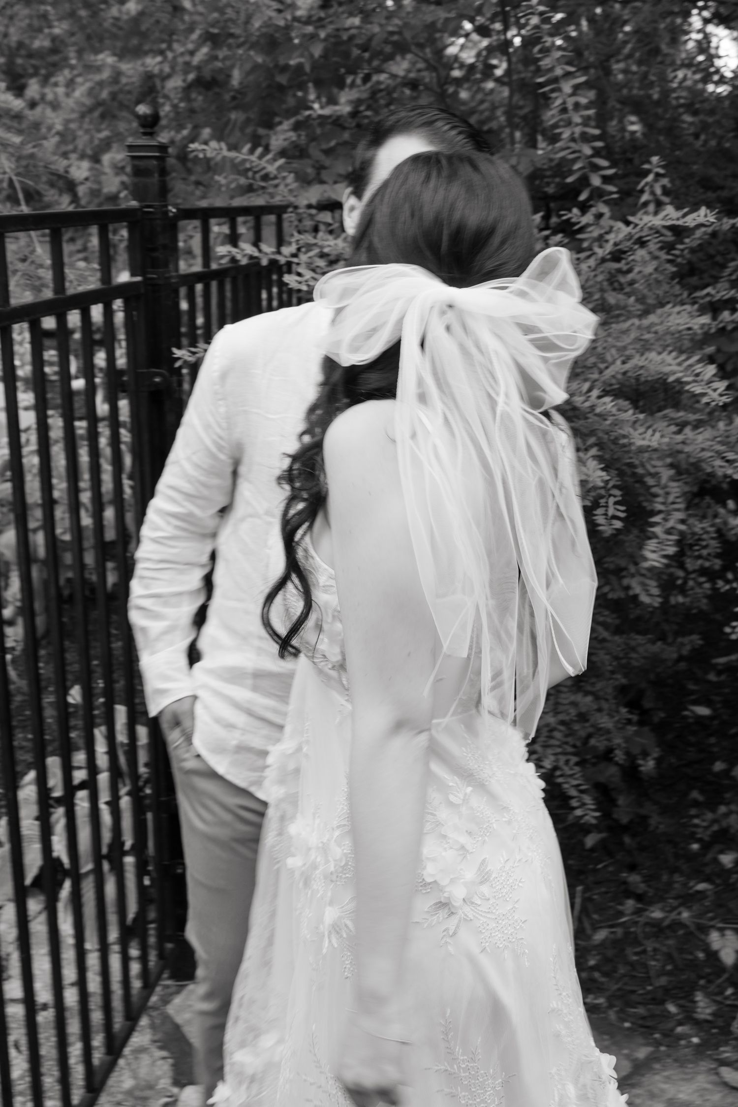 Black and white image showing the back view of a couple near an iron gate in a garden.