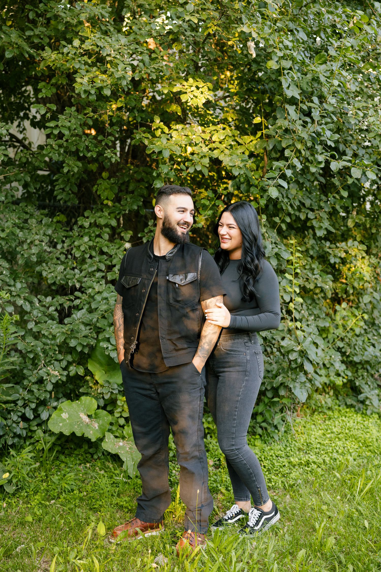 A couple shares an intimate moment while standing in front of leafy green trees in a park.