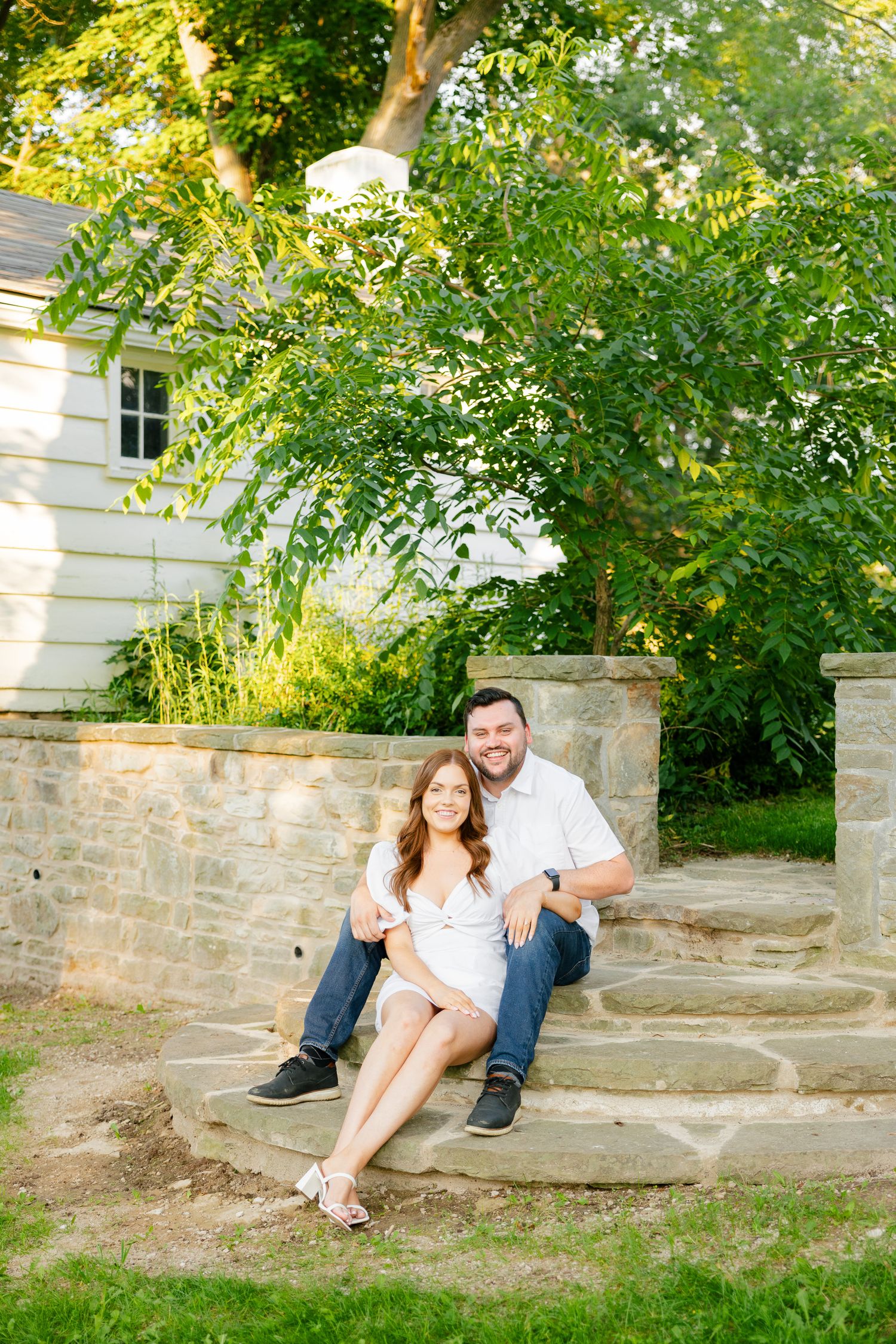 Couple relaxing on stone steps with lush greenery and white house in background.