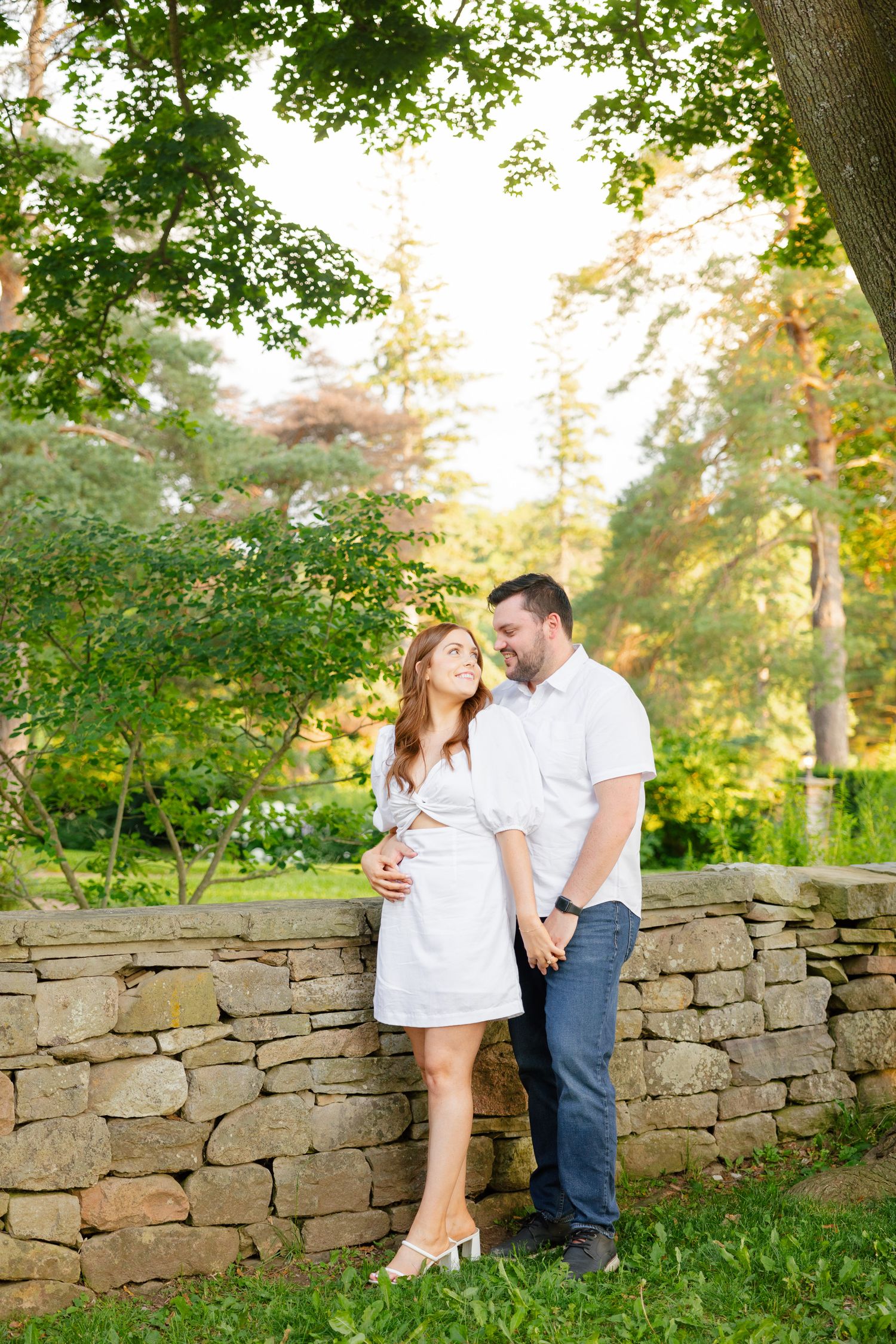 Couple in white and denim poses against a rustic stone wall in a serene garden setting.