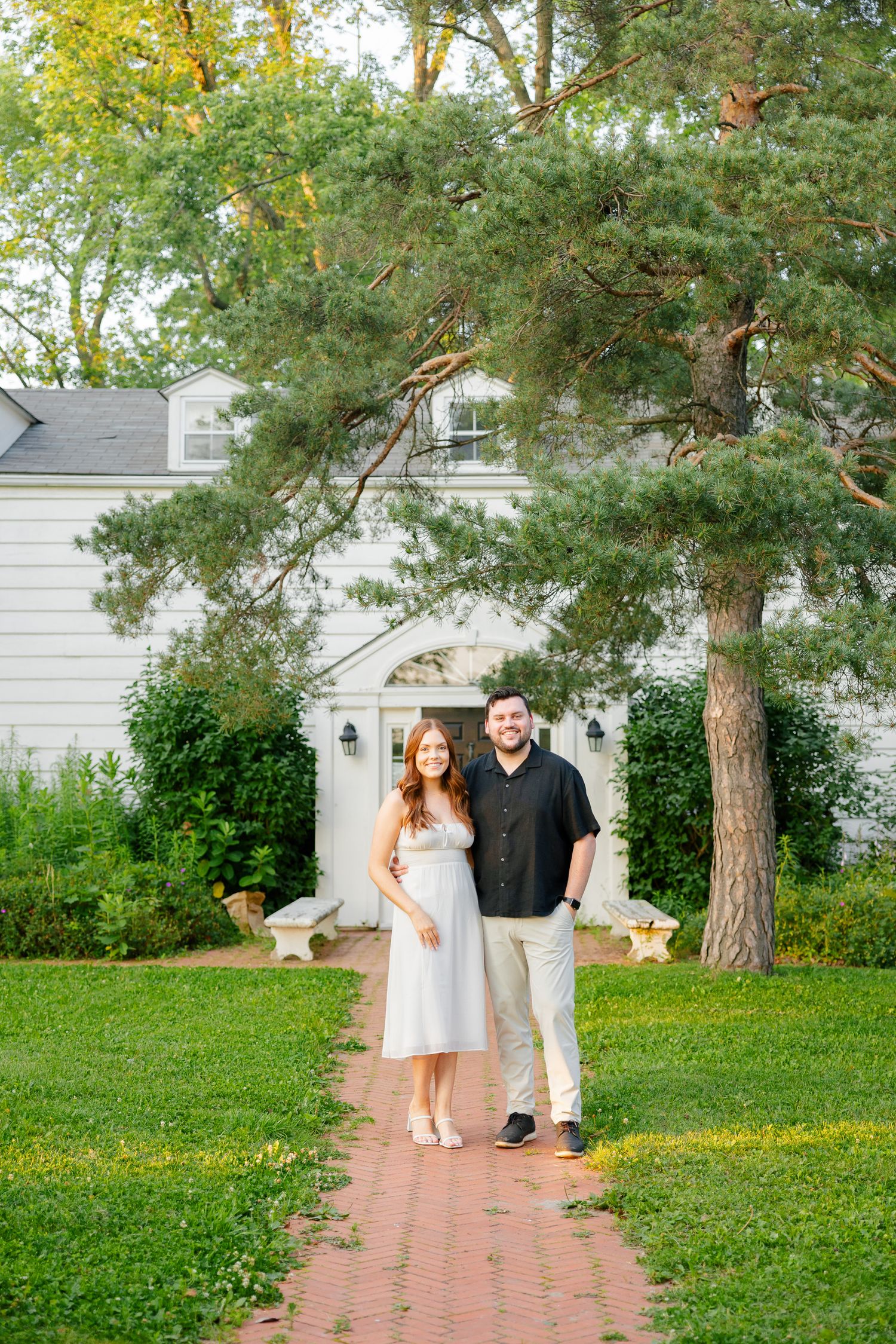 Couple standing on brick pathway leading to white house with tall pine trees overhead.