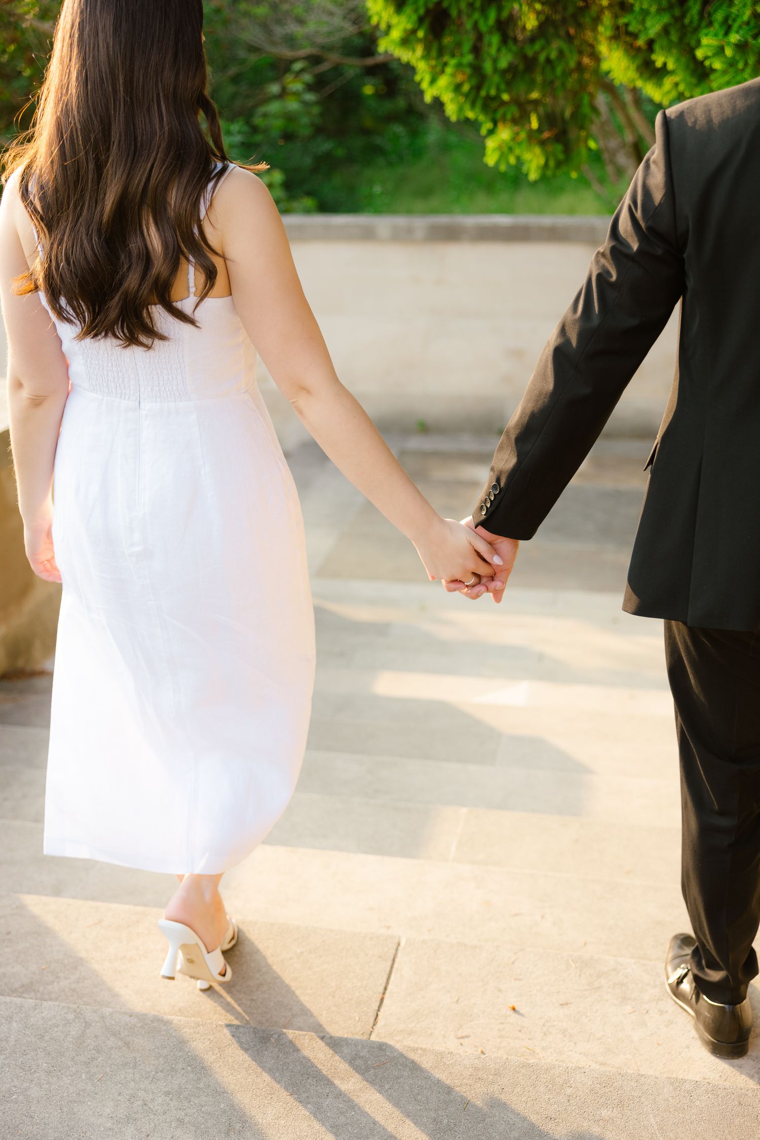 Close-up of couple holding hands while walking on stone pavement in formal attire.
