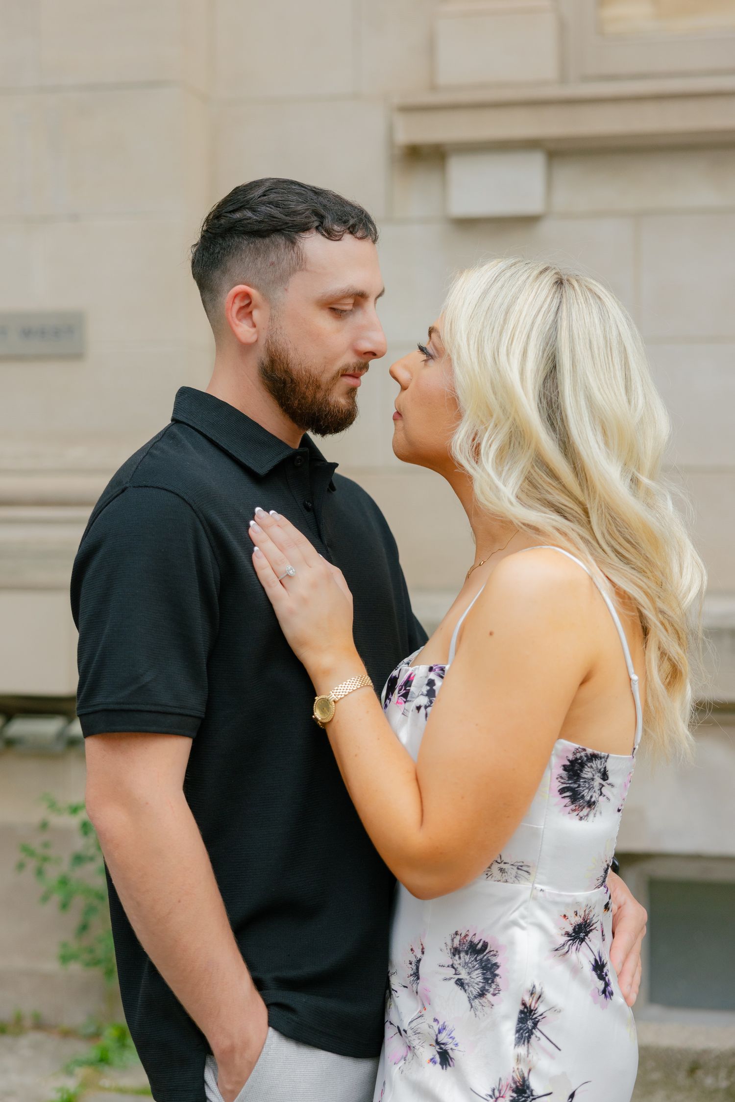 Couple in romantic pose against stone building wearing black polo and floral dress.