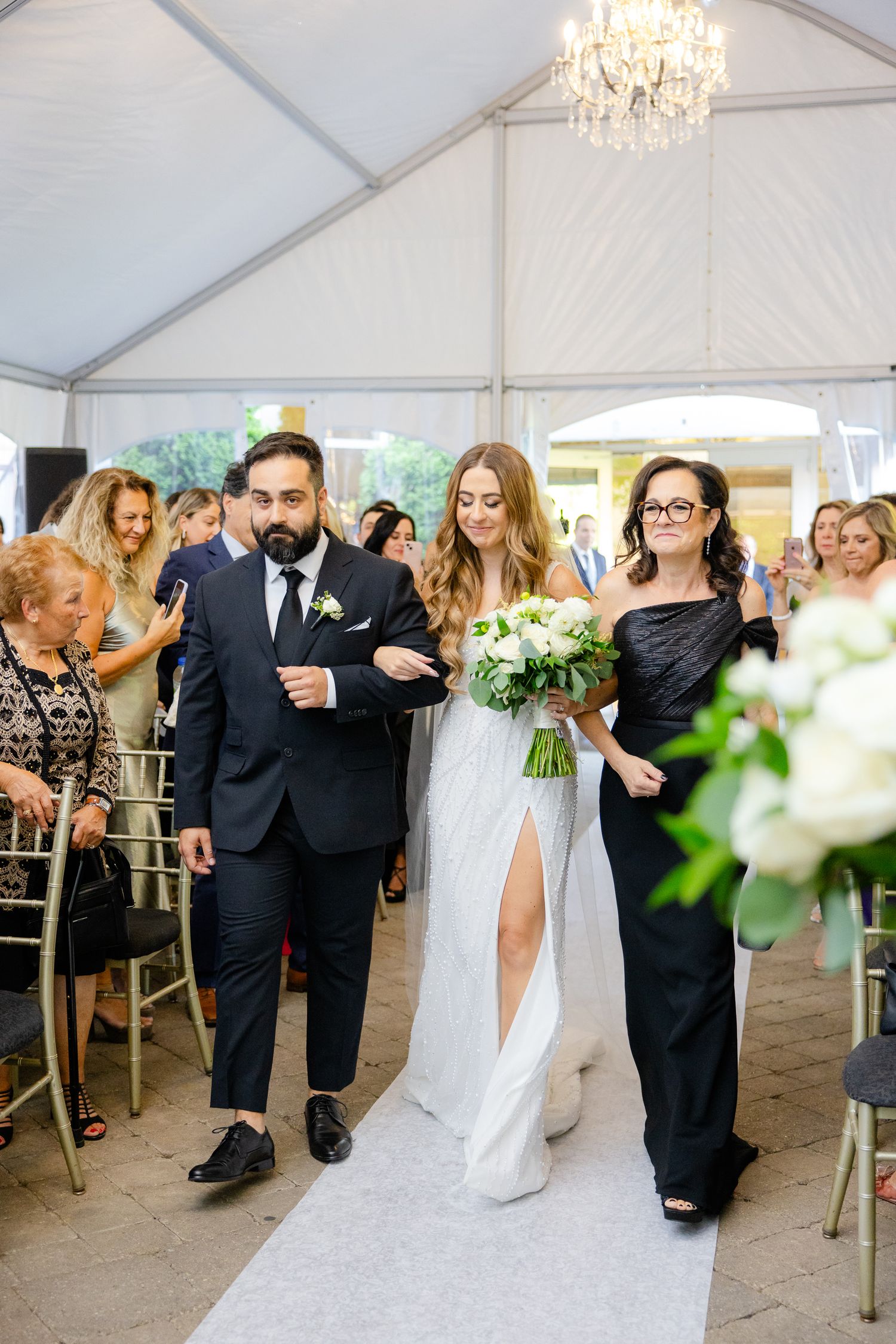 Bridal party walks down white aisle runner during wedding ceremony in elegant tented venue.
