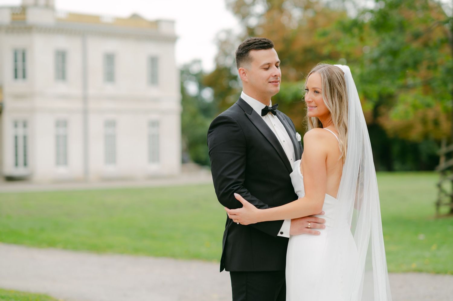 Elegant wedding couple embraces in front of classic white mansion with manicured grounds.