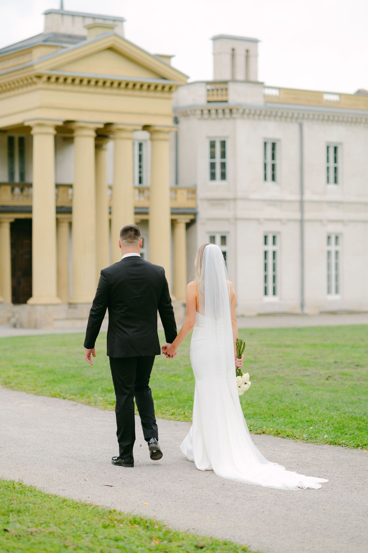 A newlywed couple walks toward a grand historic mansion with classical architecture.