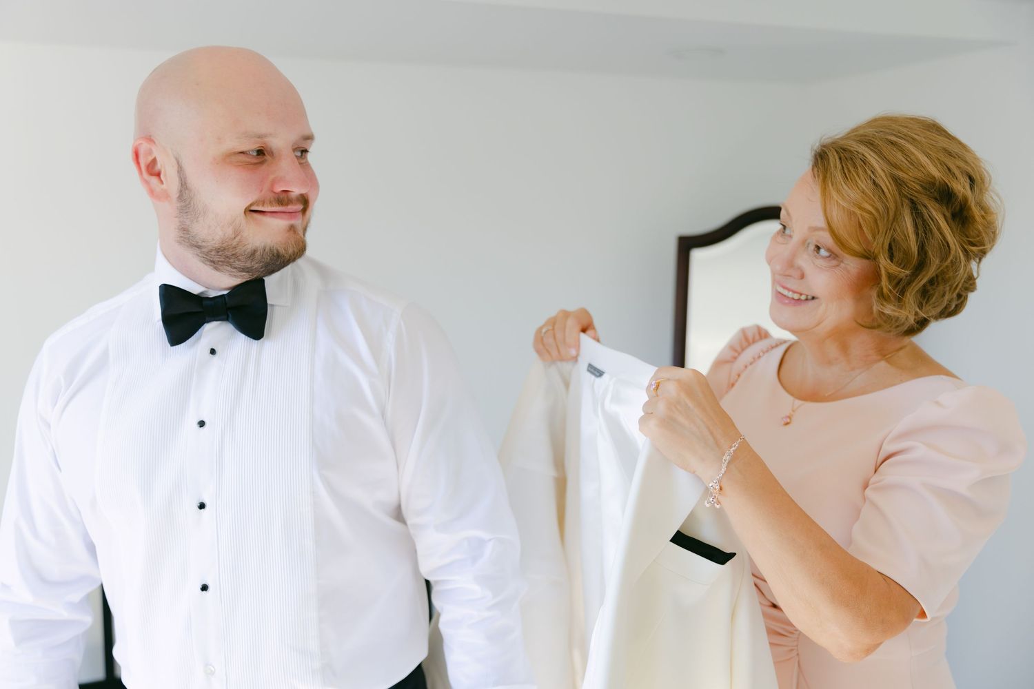 Two people in formal attire smiling during wedding preparation.