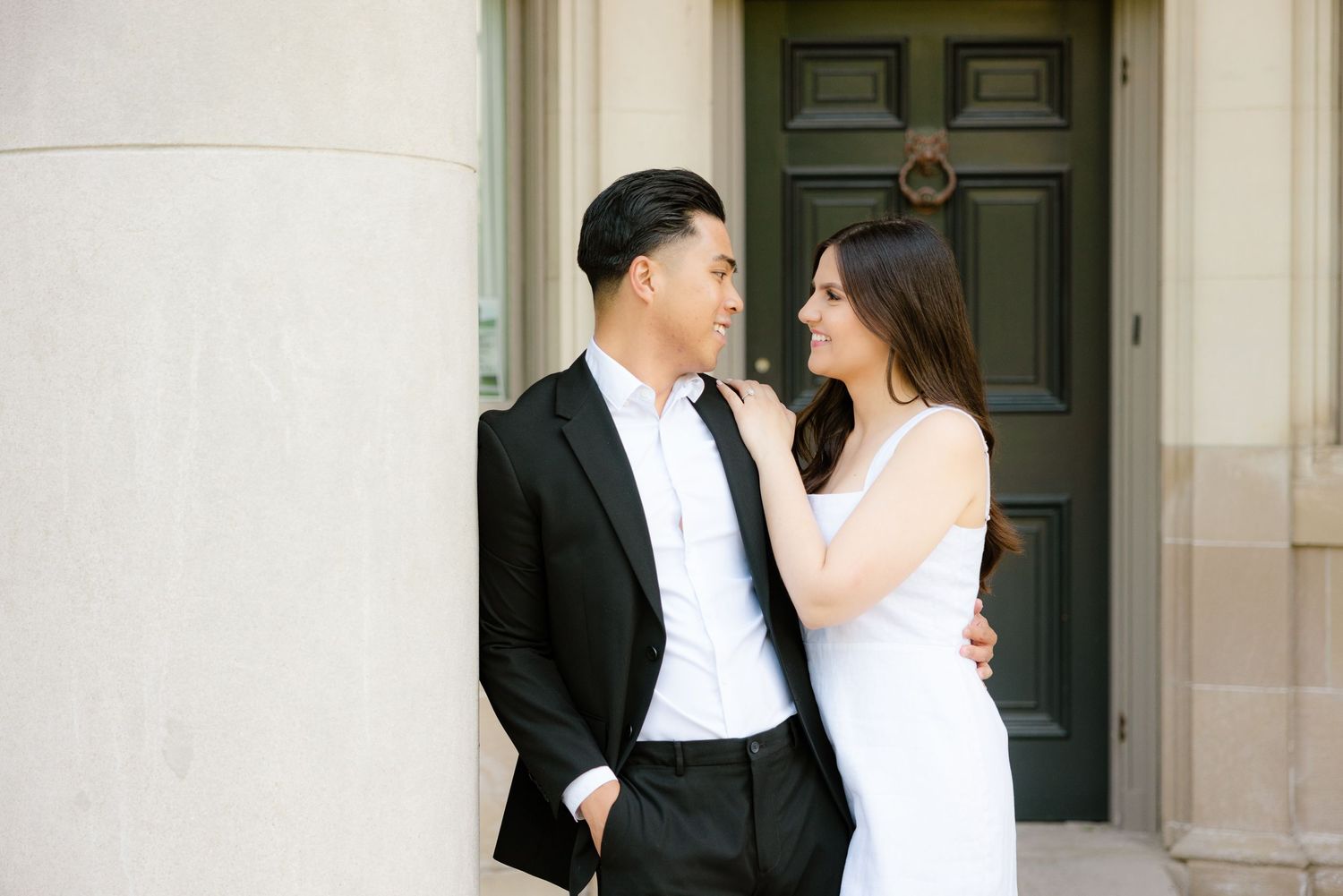 Couple in contrasting formal wear embrace while standing against cream colored building exterior.
