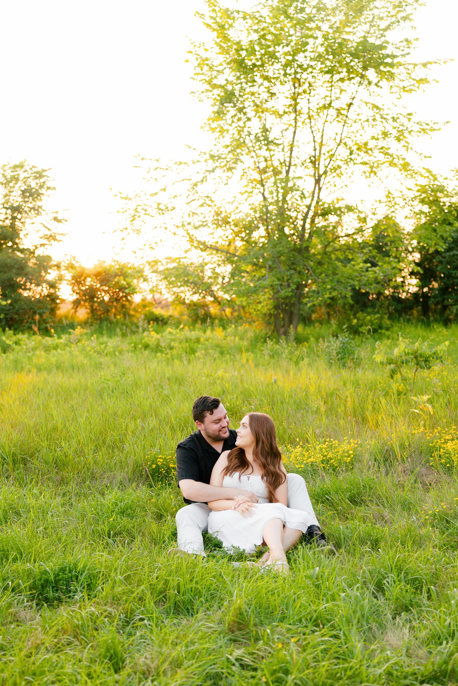 Couple shares intimate moment sitting in sunlit field with trees in background.