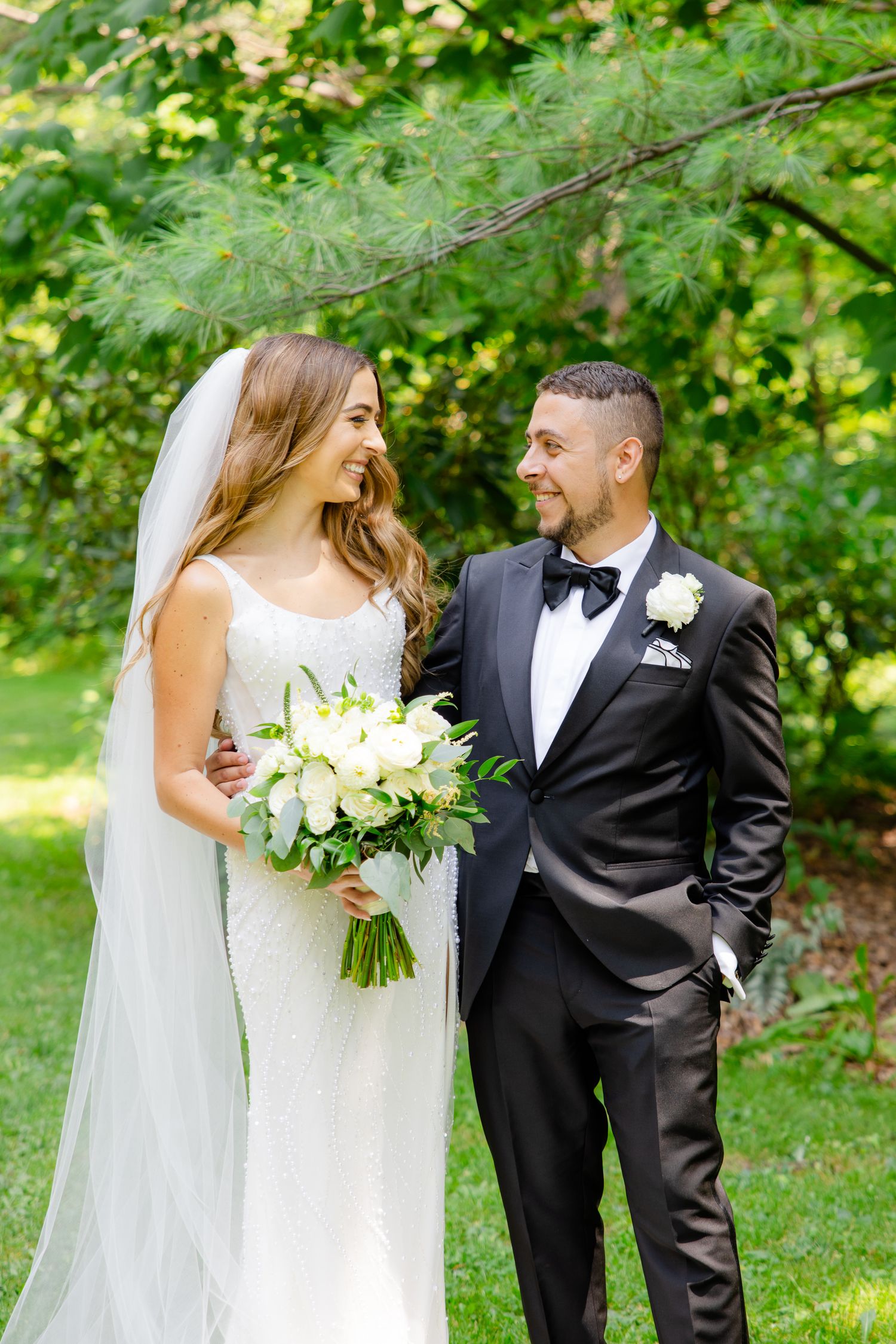 Joyful wedding portrait in a sunlit garden with white floral bouquet.