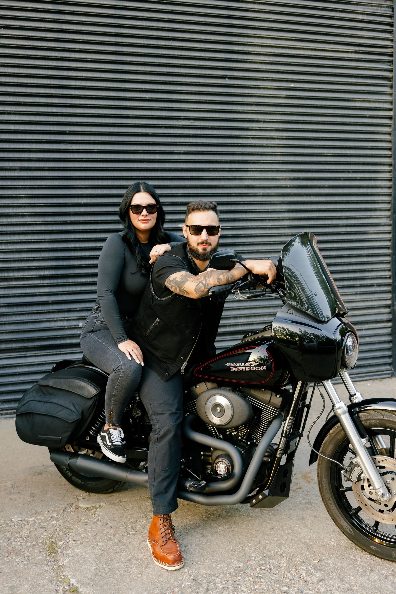 Fashionable couple in black sits on parked motorcycle near industrial building.