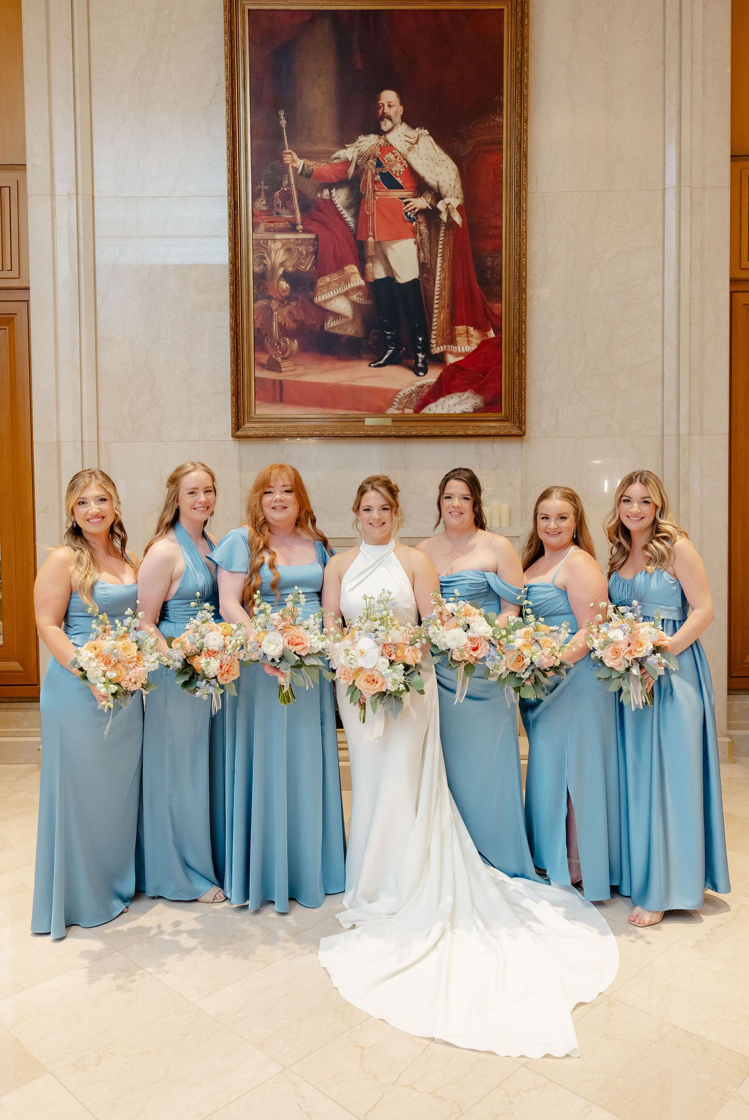 Wedding party in light blue bridesmaid dresses lined up against a formal wall with portraits.
