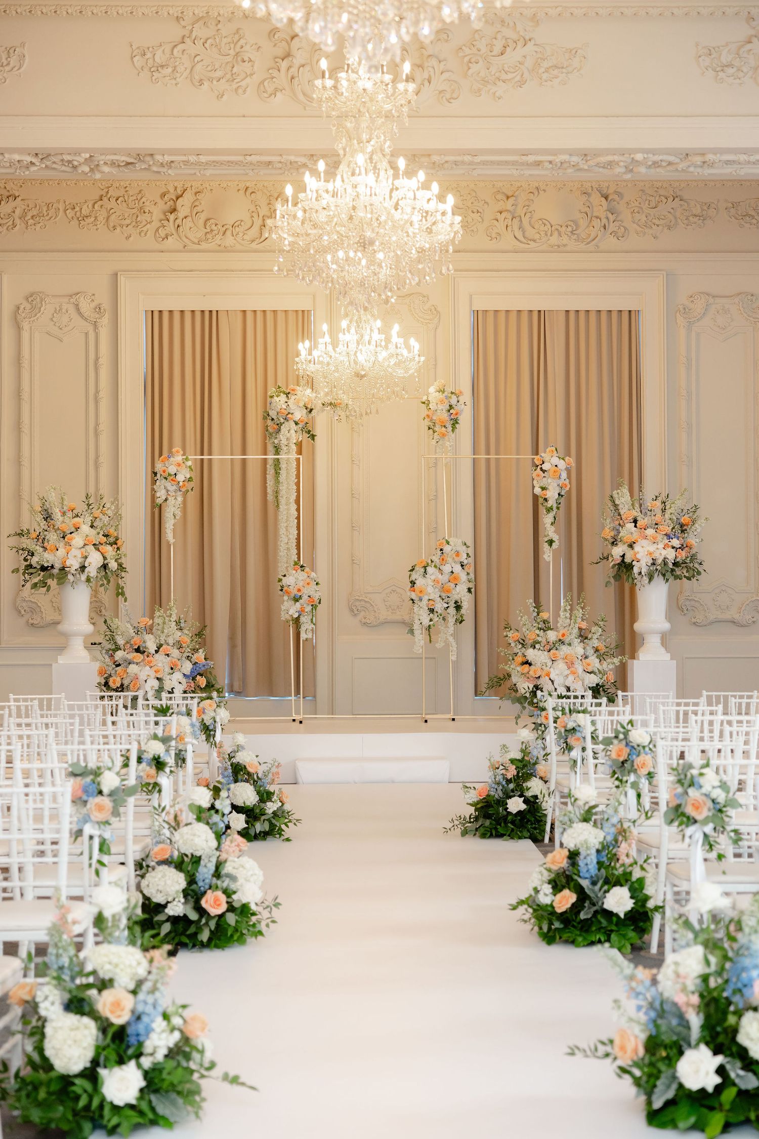 Elegant white ballroom with crystal chandeliers decorated with floral arrangements and white chairs.