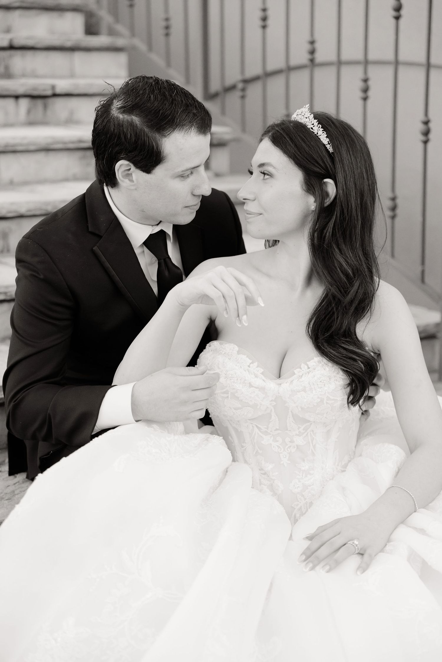 A romantic black and white wedding portrait of a couple sitting together on stairs with ornate railings.