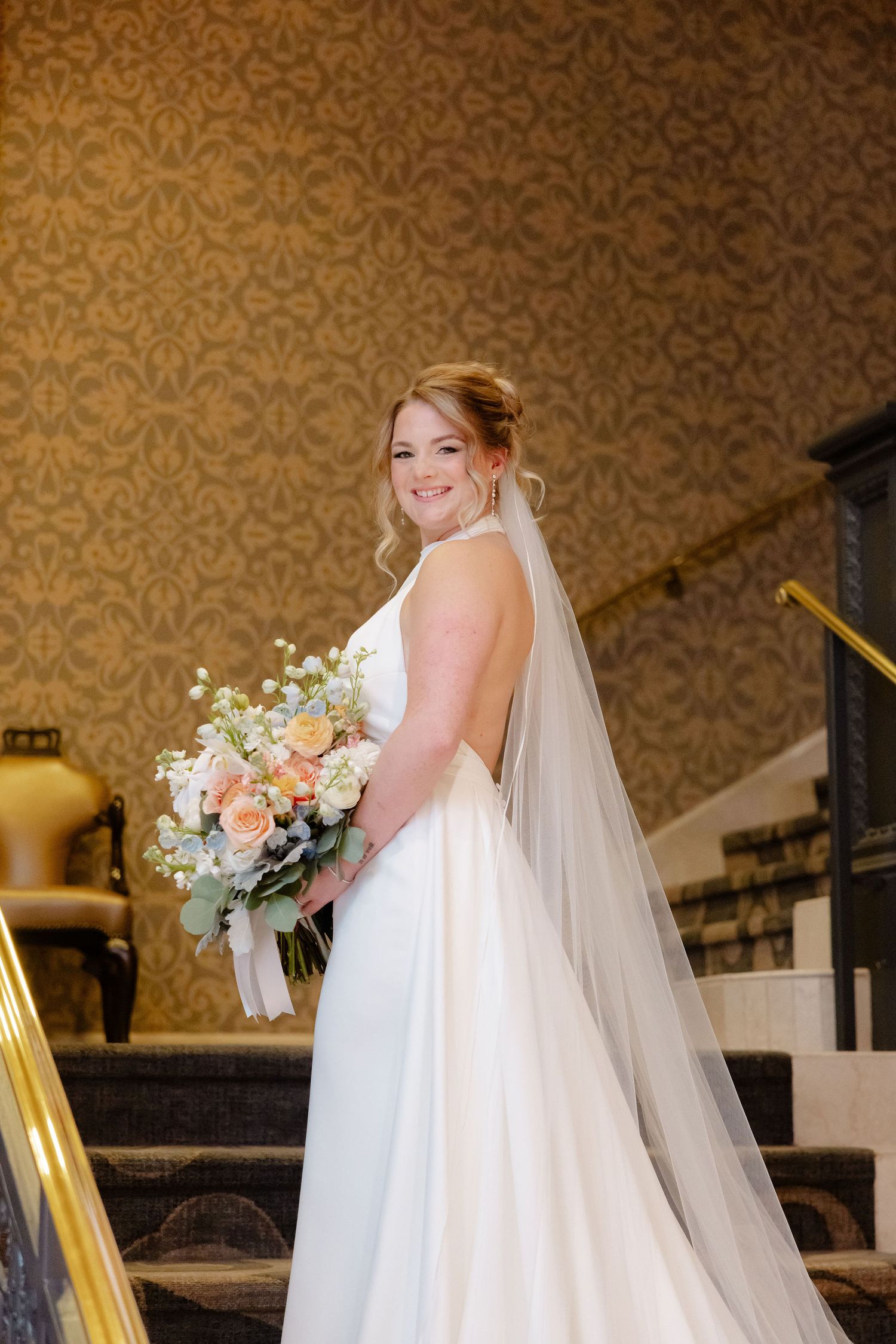 A bride in a white wedding dress and veil holds a pastel bouquet against an ornate wallpapered background.