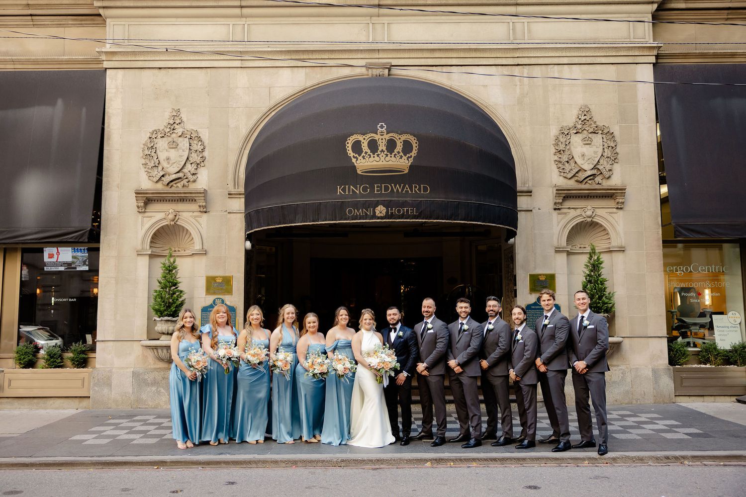 Wedding party lined up outside historic building facade for formal group portrait.