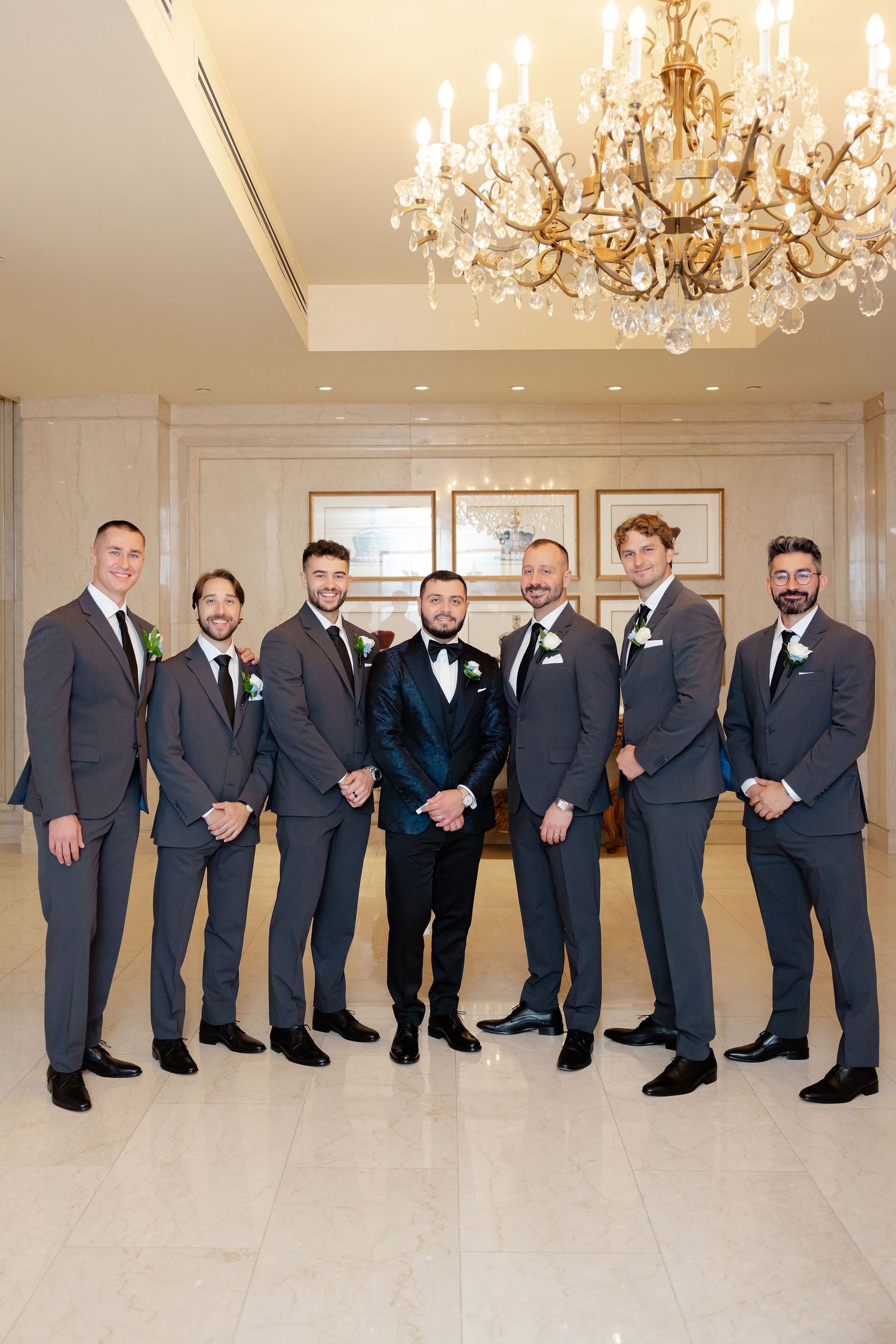 Wedding party members in dark suits lined up under crystal chandeliers in an elegant ballroom.