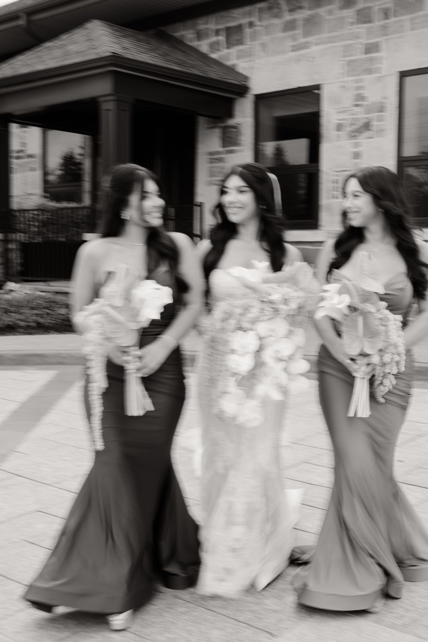 Wedding party in dark dresses and white dress walking and holding bouquets at stone building entrance.