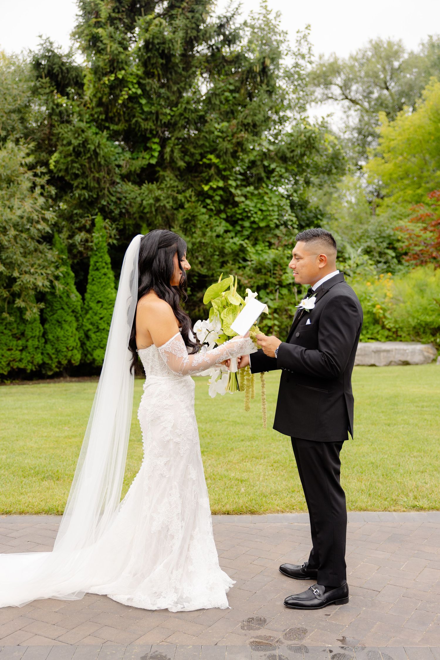 A couple in wedding attire shares a playful dance sequence outdoors on a paved area surrounded by green trees.