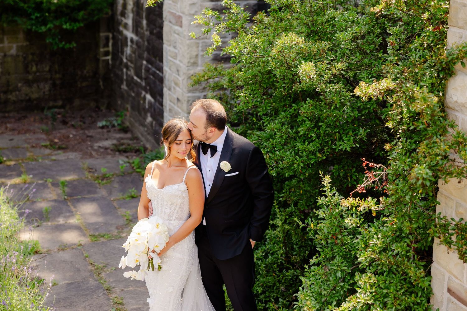 Bride and groom embrace in keyhole garden archway with blooming flowers and stone walls providing a romantic backdrop.