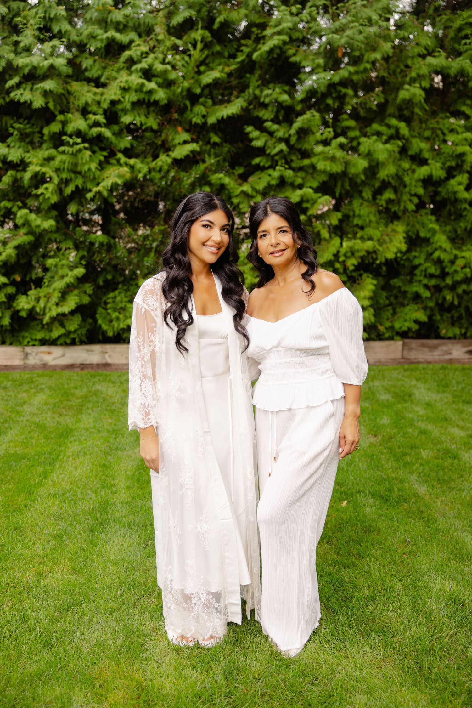Two people in white dresses stand together on green grass with lush foliage in the background.