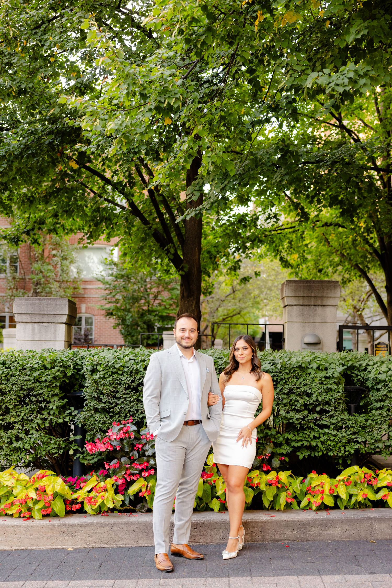 A couple poses together on a brick path surrounded by manicured hedges and vibrant flowers.