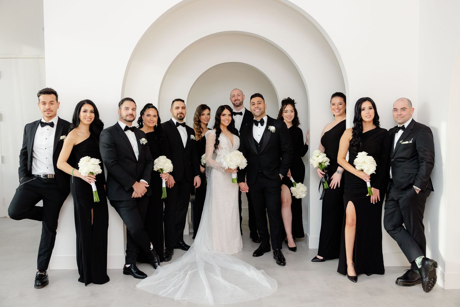 Wedding party in formal black and white attire poses under modern white archway.