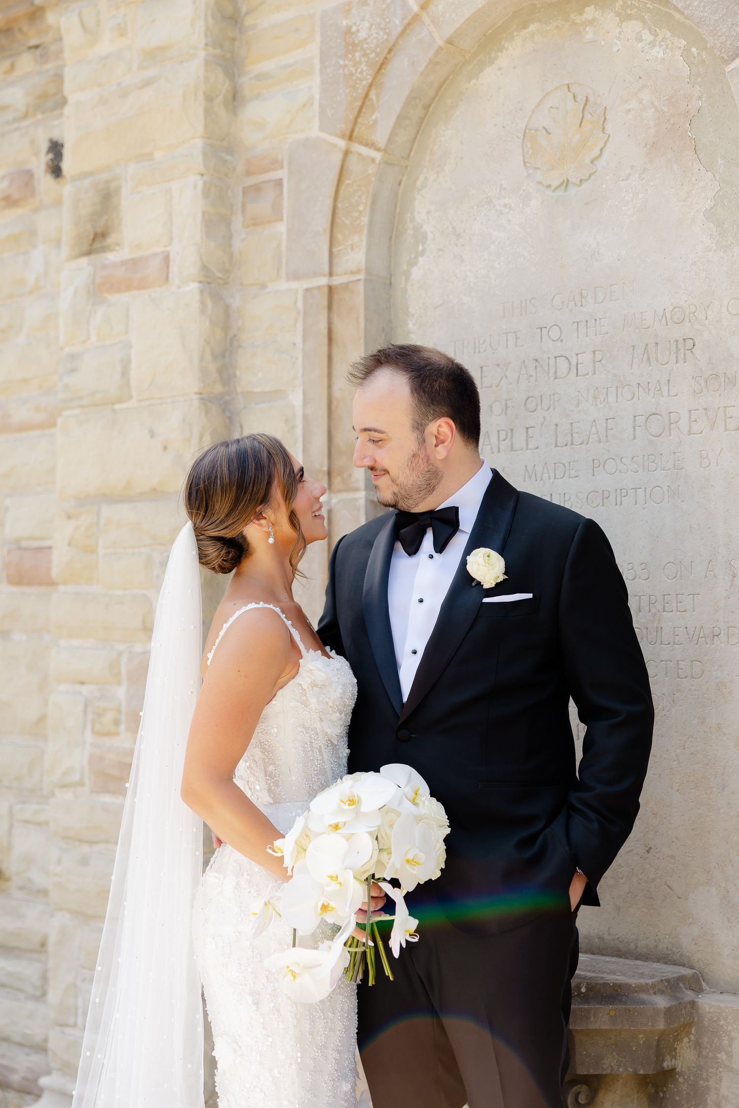 A wedding couple stands intimately together against a stone church wall with the bride holding a white floral bouquet.
