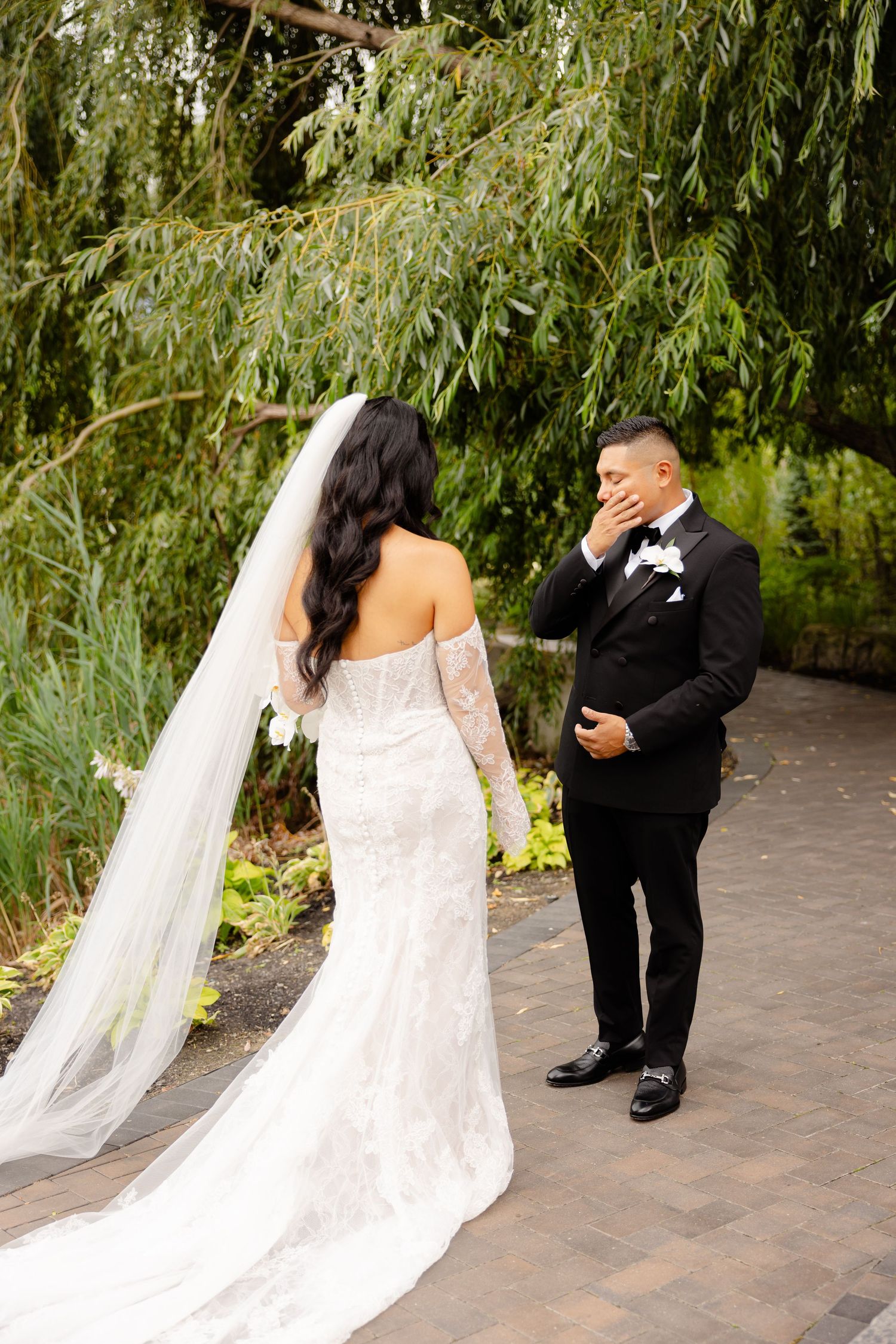 Bride and groom share first look moment under weeping willow trees in garden setting.