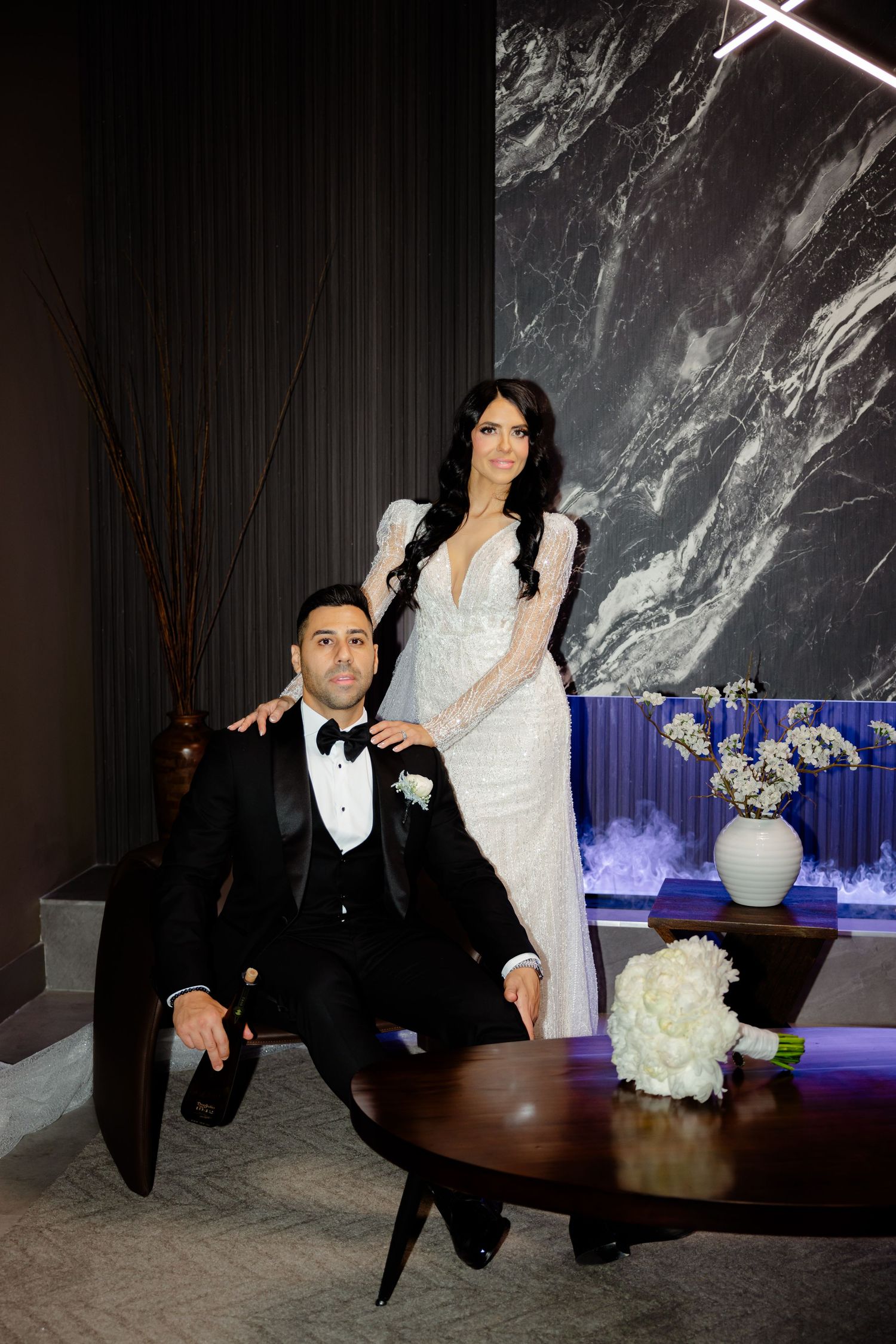 Wedding couple poses by decorative table with white floral bouquet and marble wall backdrop.