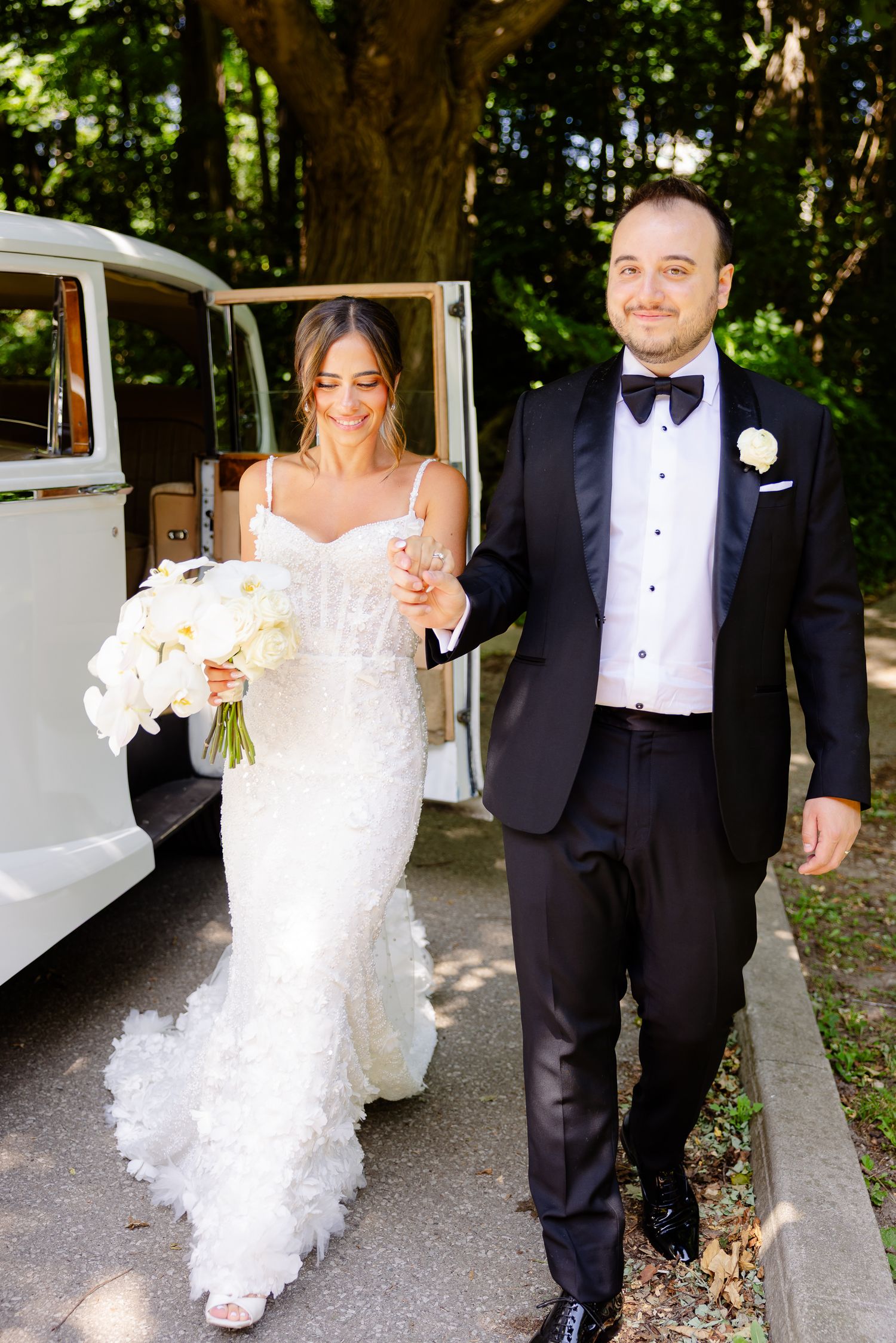 A bride in a lace mermaid gown holding white orchids walks with groom in tuxedo next to vintage white car.