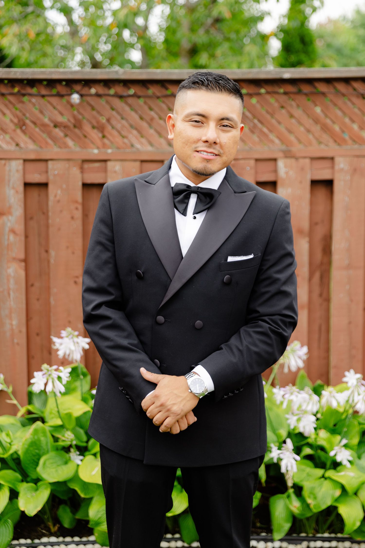 Person in black tuxedo with bowtie poses in front of wooden fence and white flowers in garden setting.