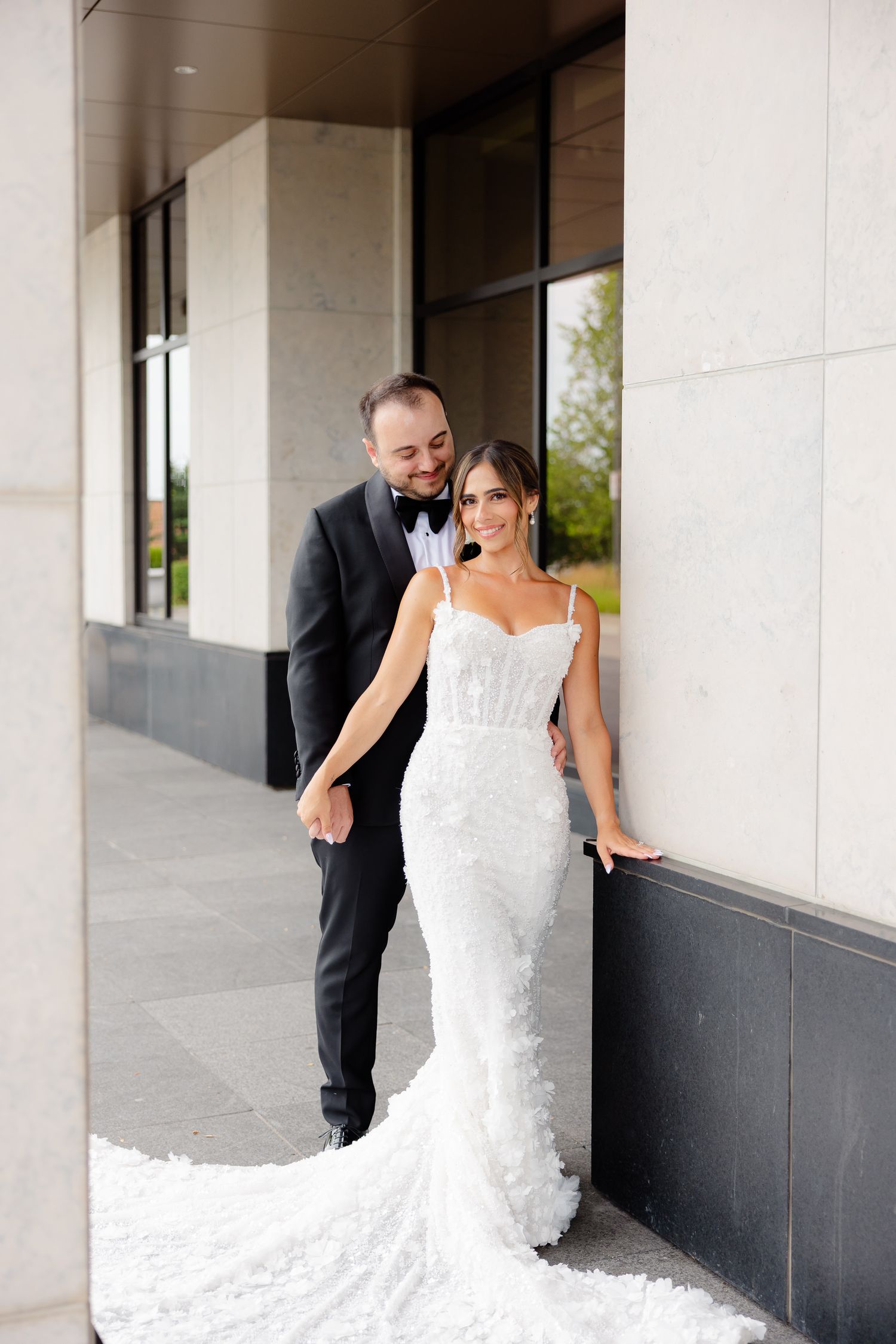 A couple poses together on their wedding day wearing formal attire with a white lace gown and black tuxedo.