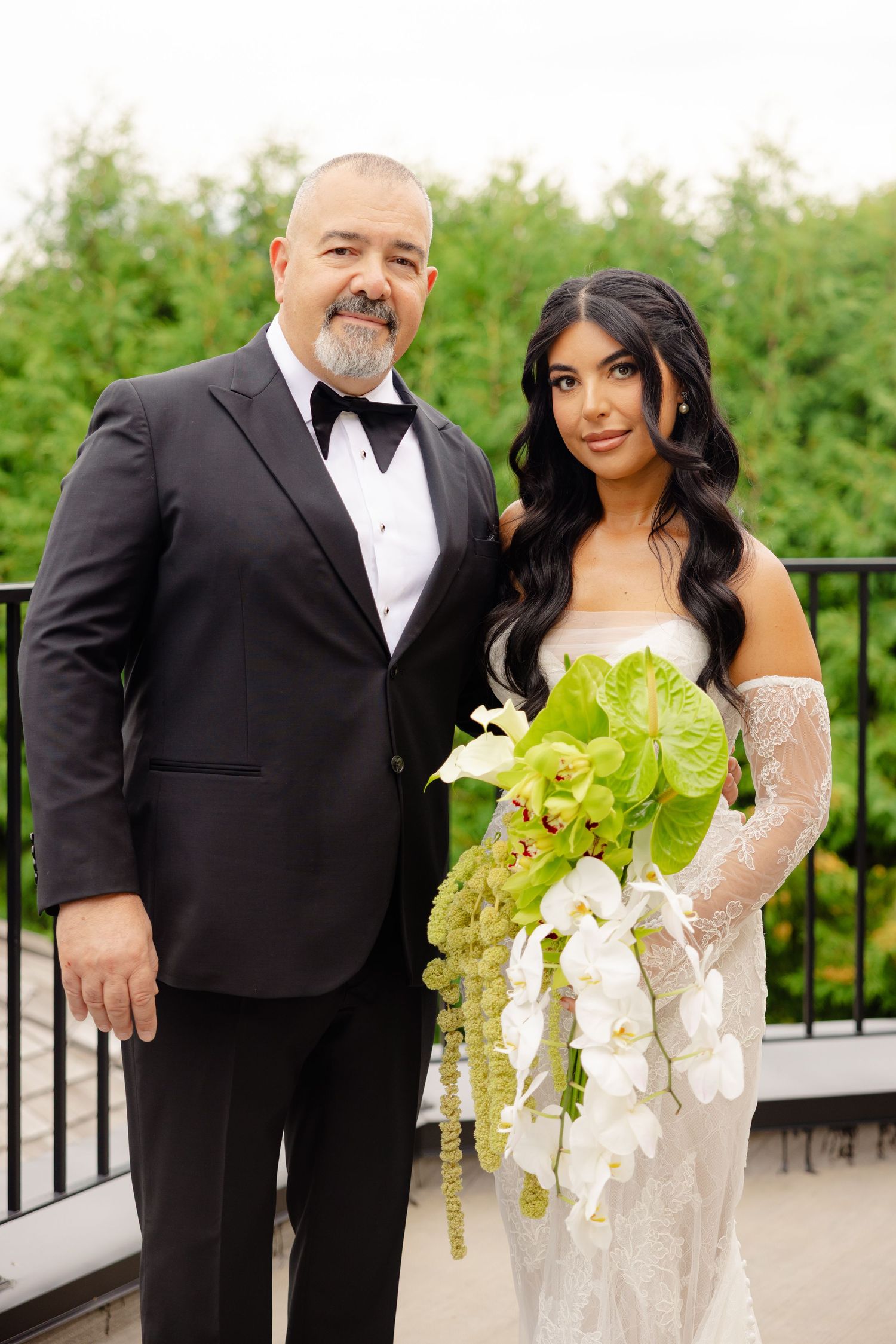 A couple in formal wedding attire poses together on an outdoor terrace with lush greenery in the background.