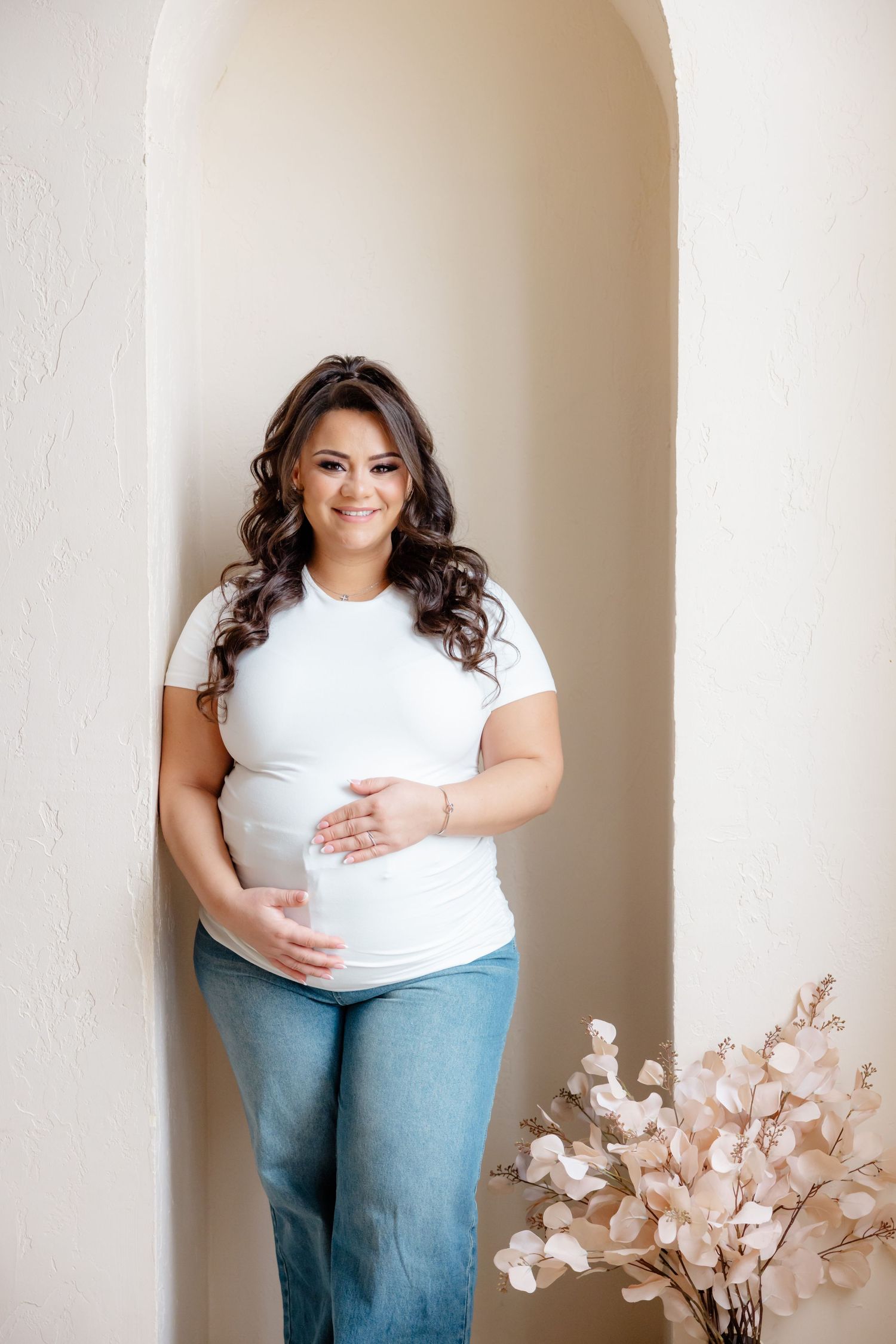 Expectant mother in white shirt and jeans stands in doorway archway next to decorative floral arrangement.