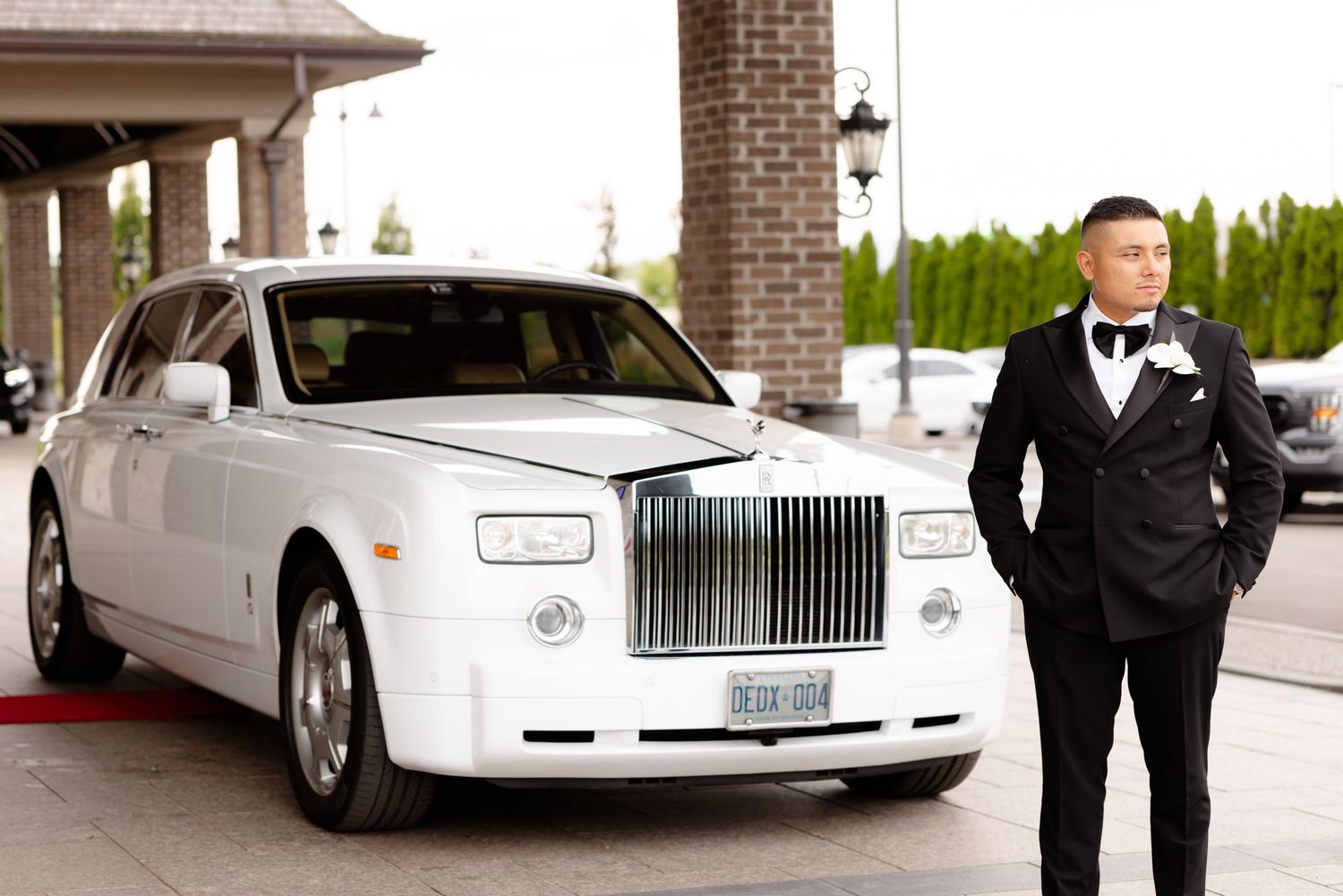 A luxury white Rolls-Royce wedding car parked in front of a brick building with elegantly dressed figures nearby.
