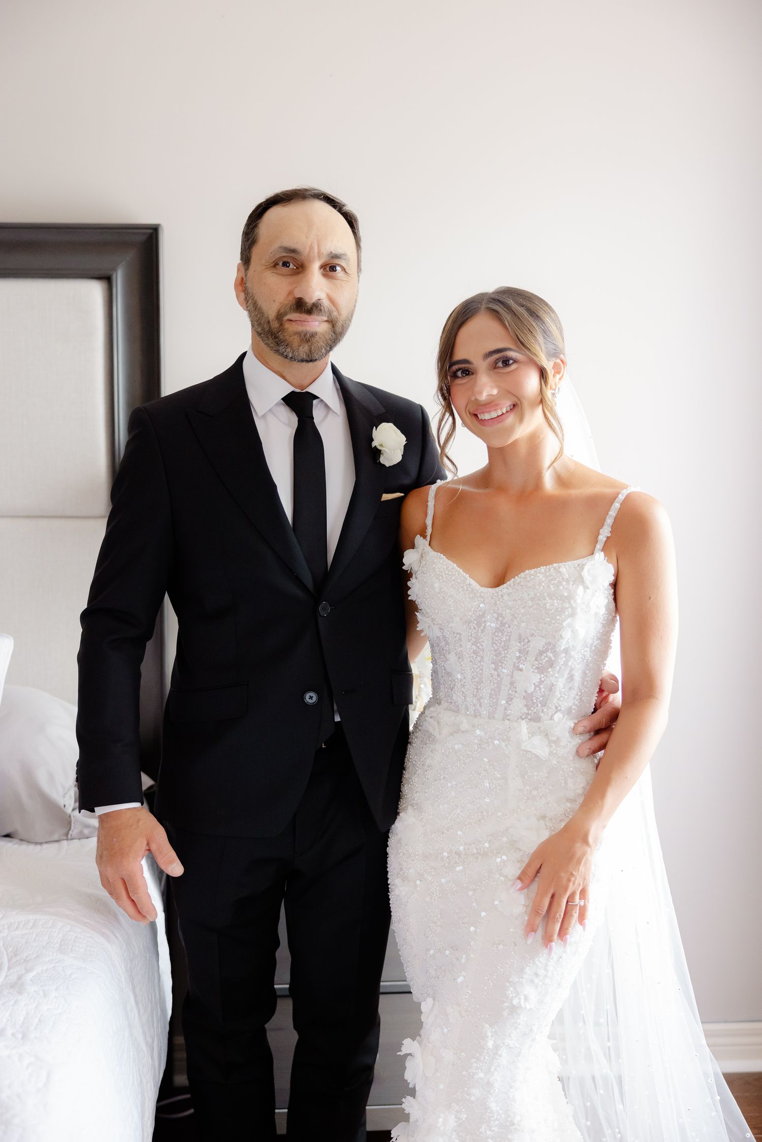 A bride in a lace spaghetti strap gown and groom in classic black suit stand side by side in a bright room.