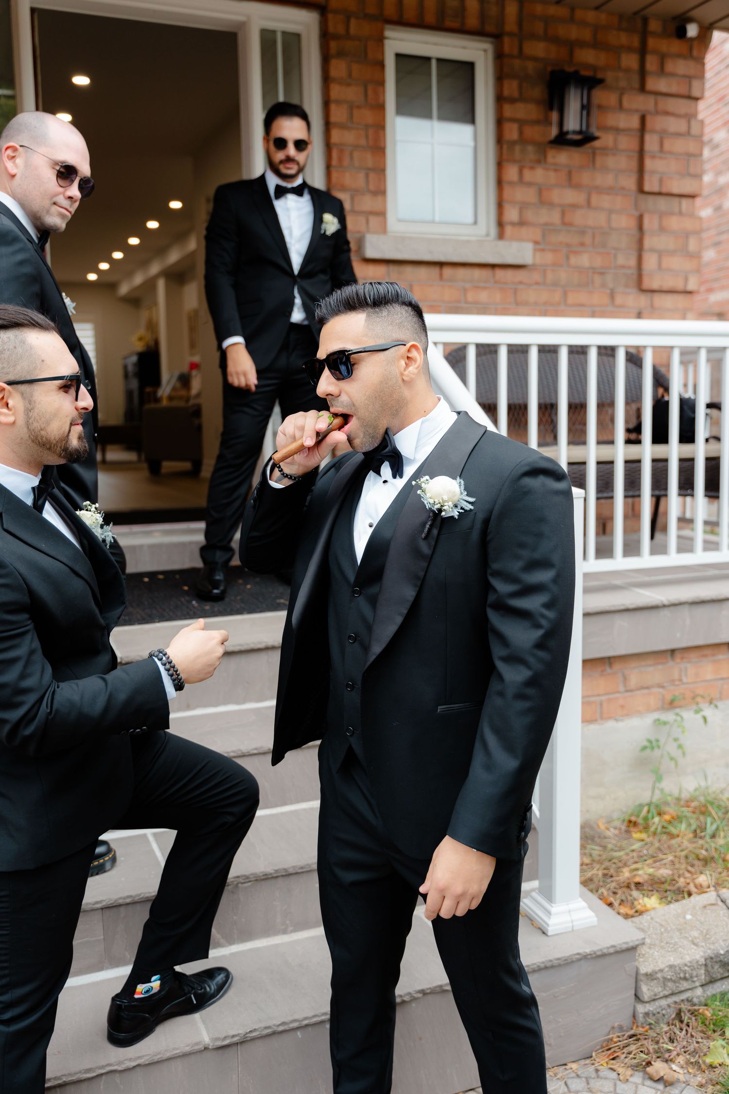 Wedding party in formal black suits and sunglasses sharing a laughing moment on porch steps.
