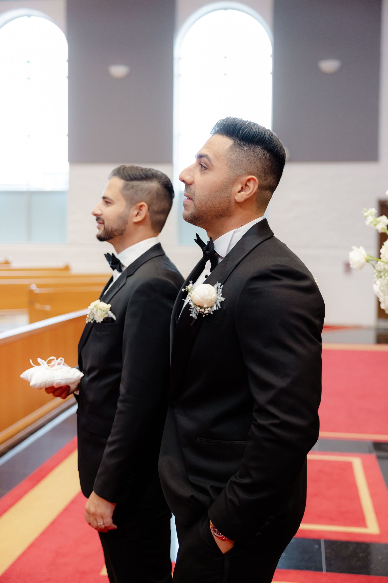 Two people in black tuxedos with boutonnieres stand at the altar during a wedding ceremony.