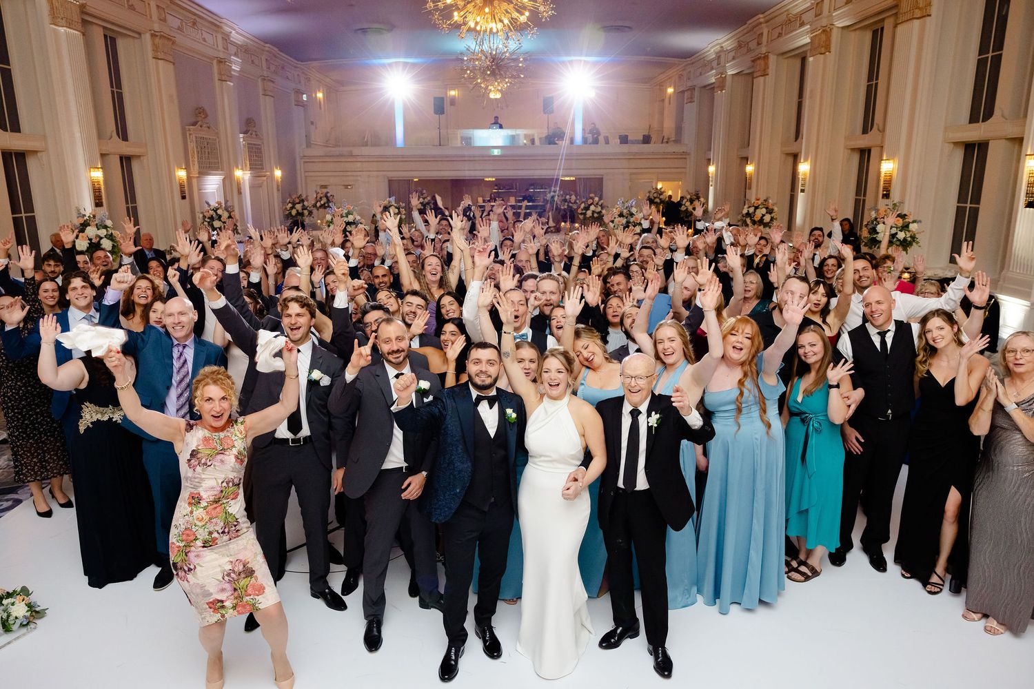 Large wedding reception crowd celebrating with raised hands in an ornate ballroom.