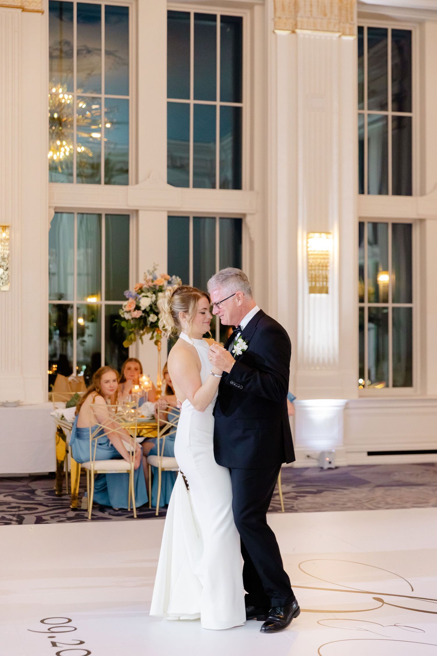 A couple shares their first dance in an elegant ballroom with tall windows and floral decorations.