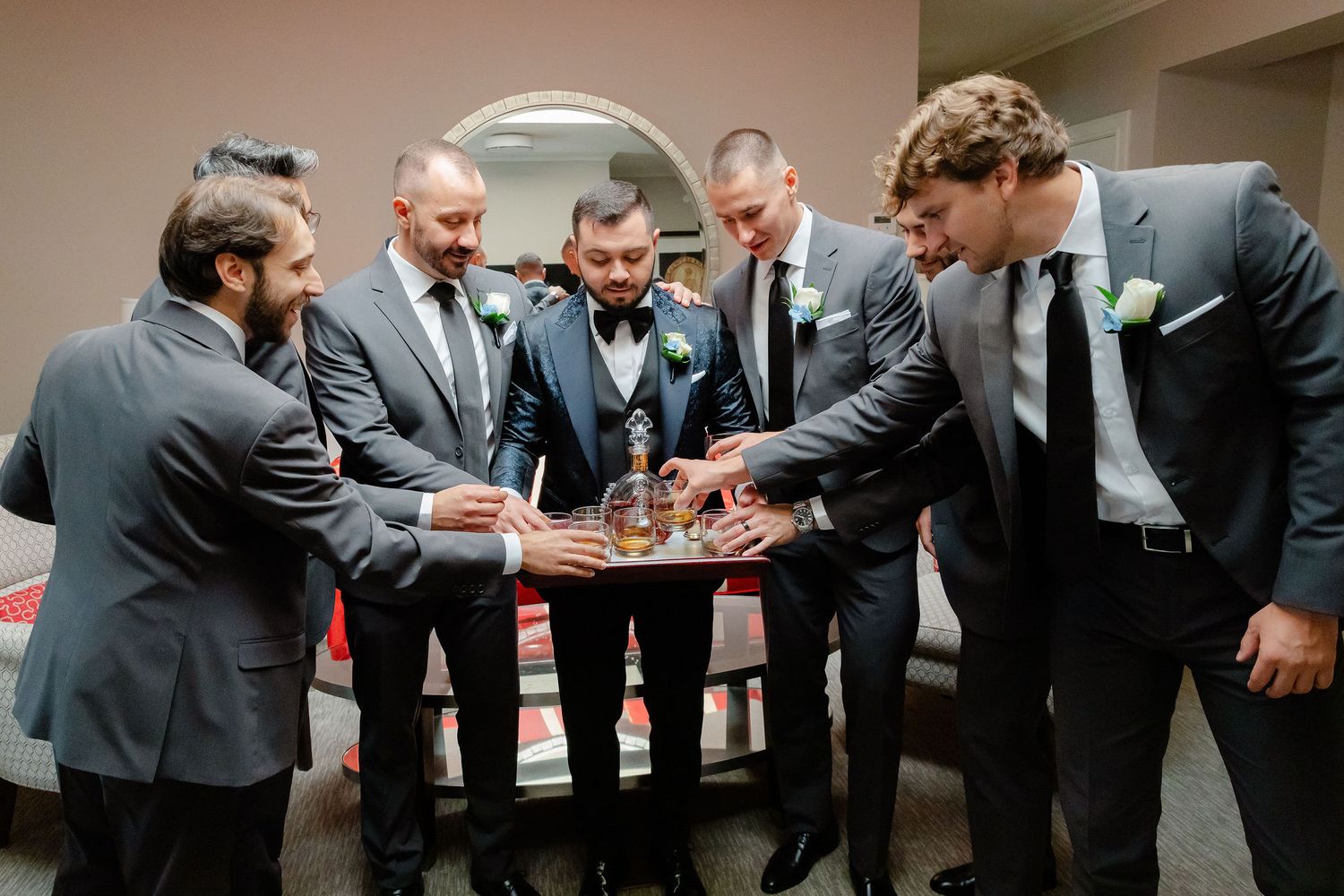 A group of groomsmen in dark suits gather in a circle for a pre-wedding prayer moment.
