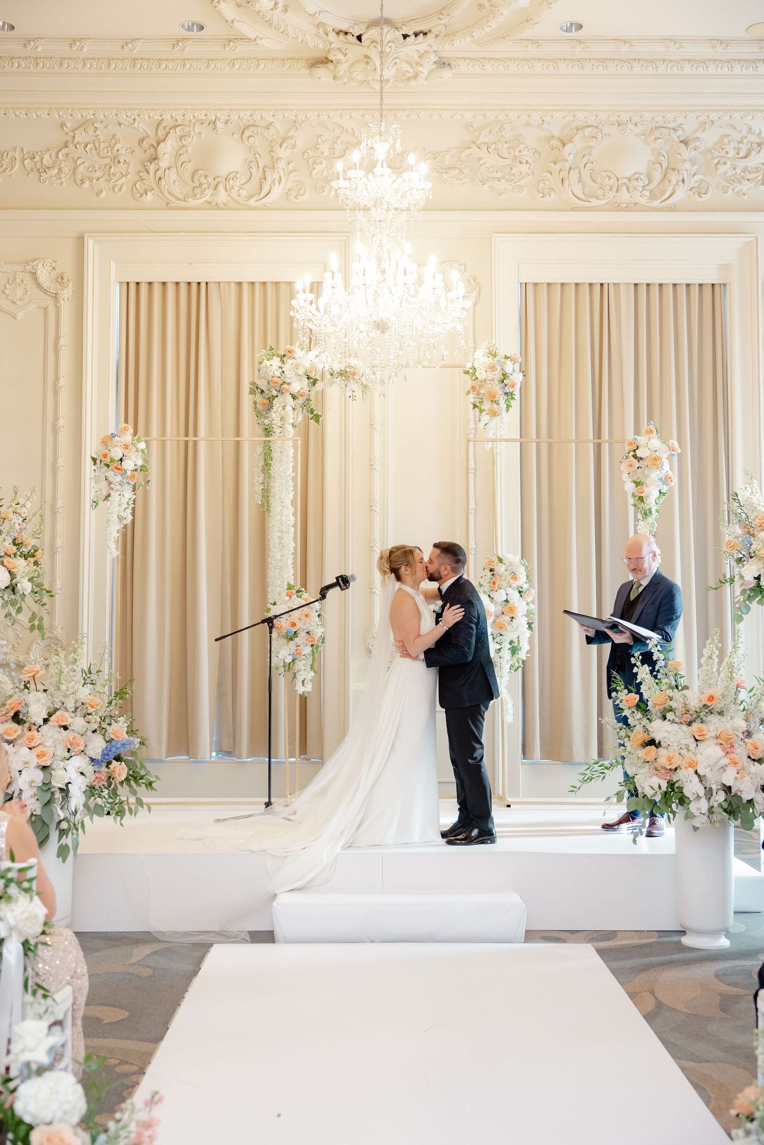 Wedding ceremony moments captured in elegant white ballroom with floral arrangements.